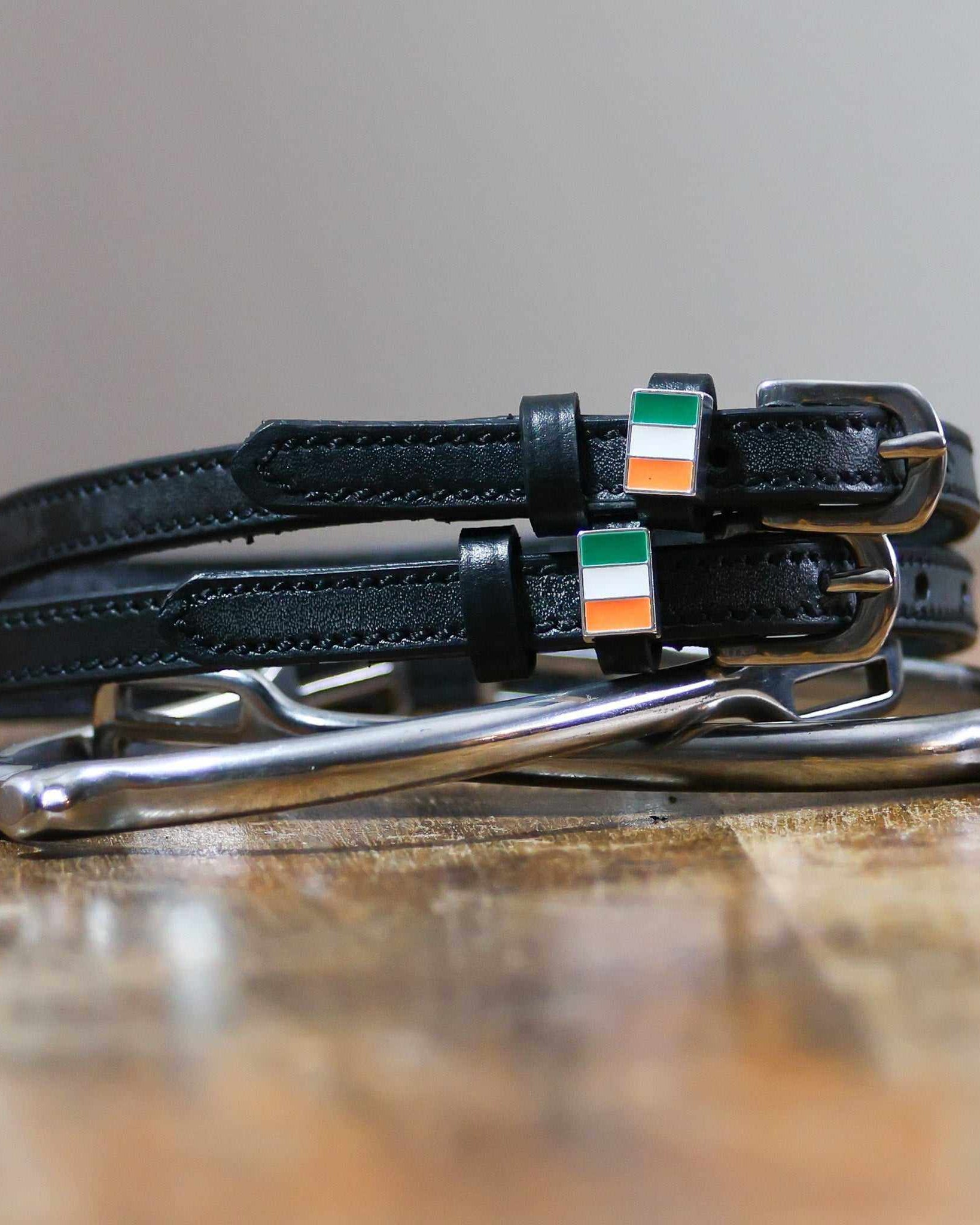 Black leather Ireland flag spur straps coiled beside a stirrup on wood