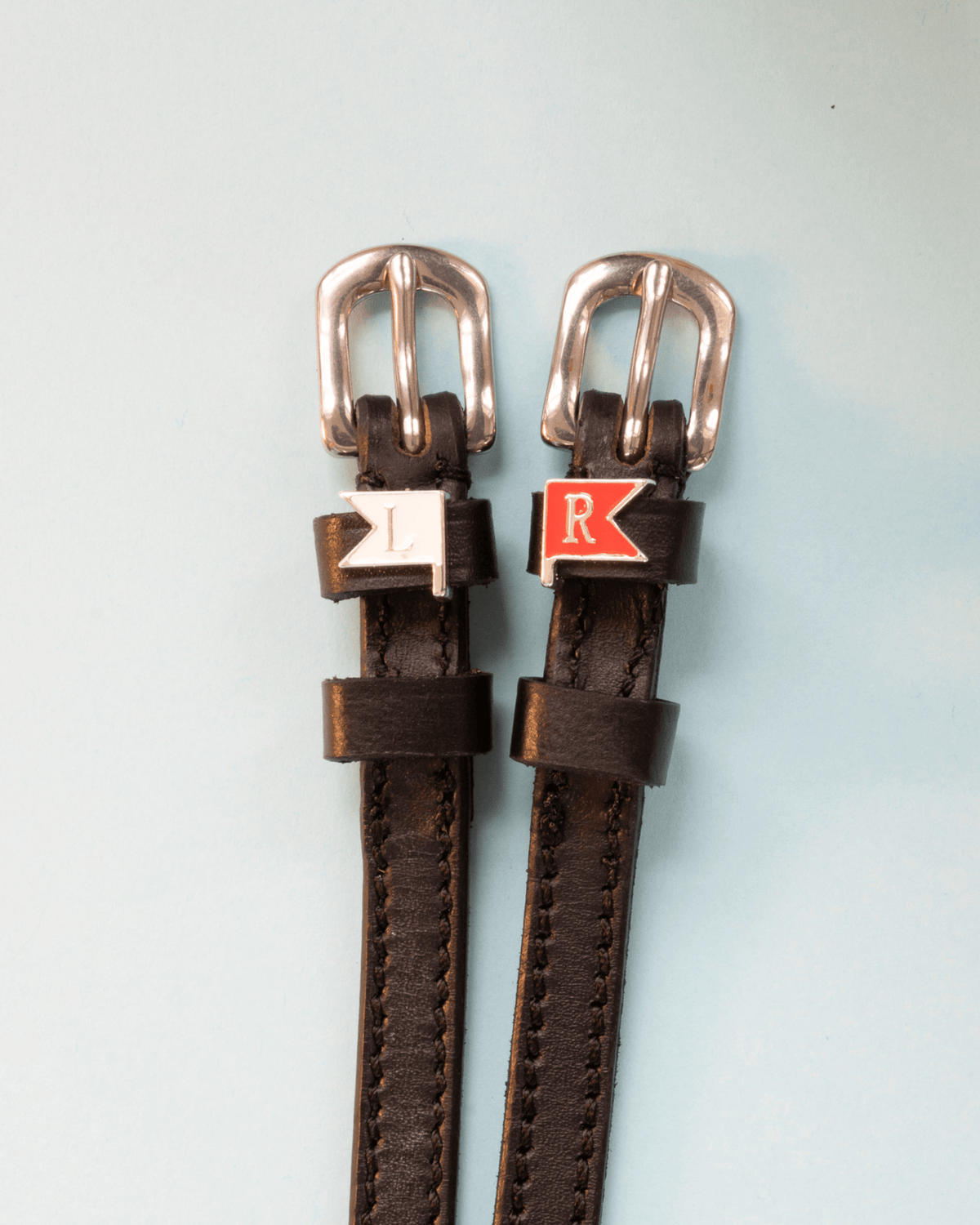 Brown leather ManeJane jump-flag spur straps with rose-gold buckles on pale blue