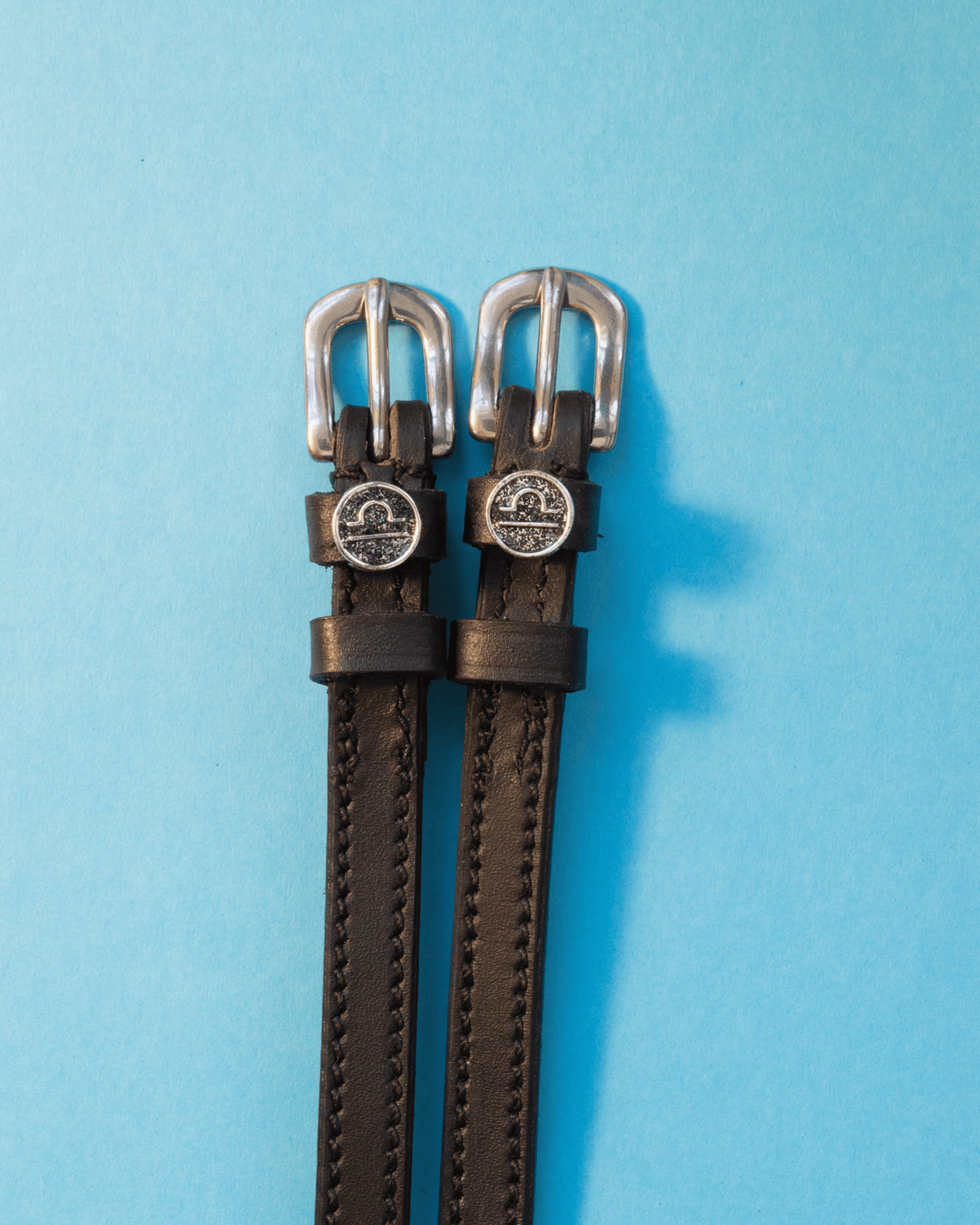 Black leather ManeJane spur straps with round Libra zodiac charms on a bright blue backdrop