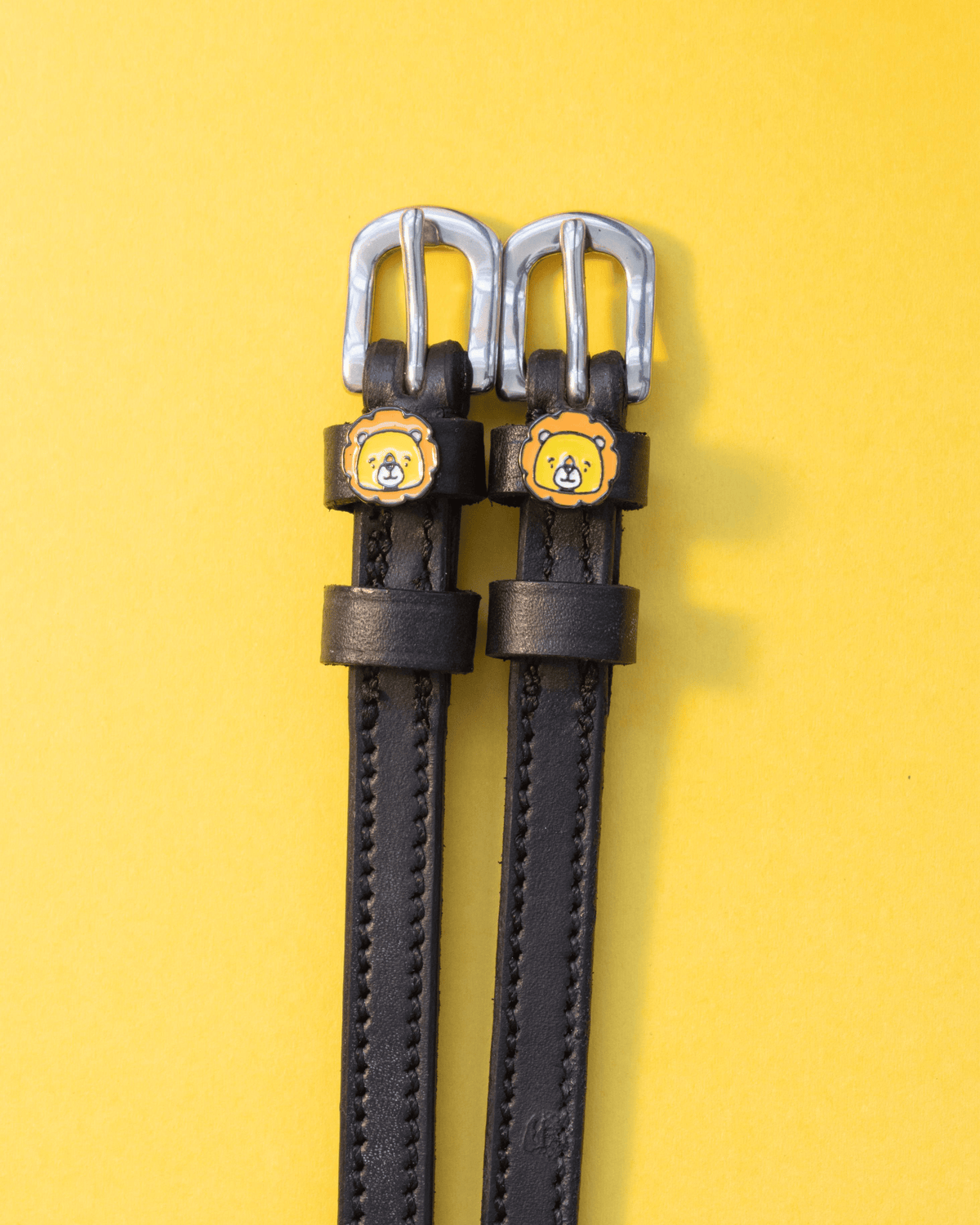 Black leather ManeJane spur straps with cartoon lion face charms and silver buckles on yellow