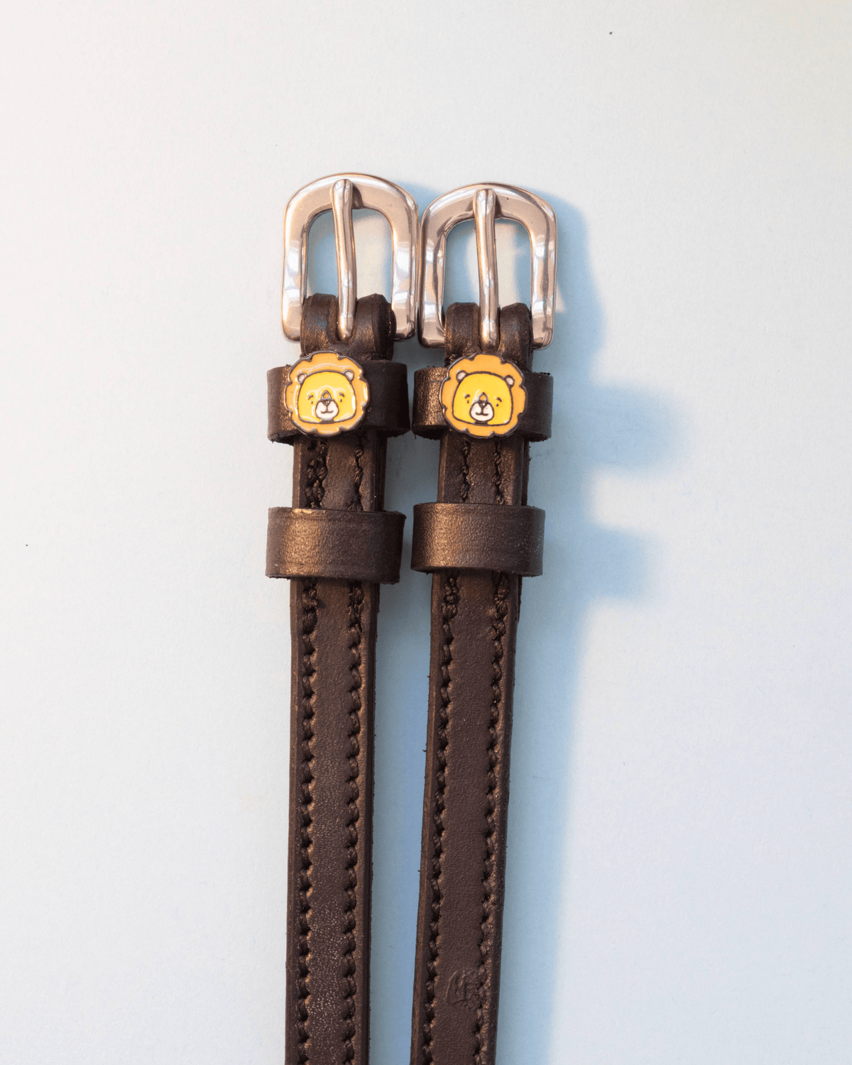 Dark brown leather lion charm spur straps with silver buckles on a pale grey background