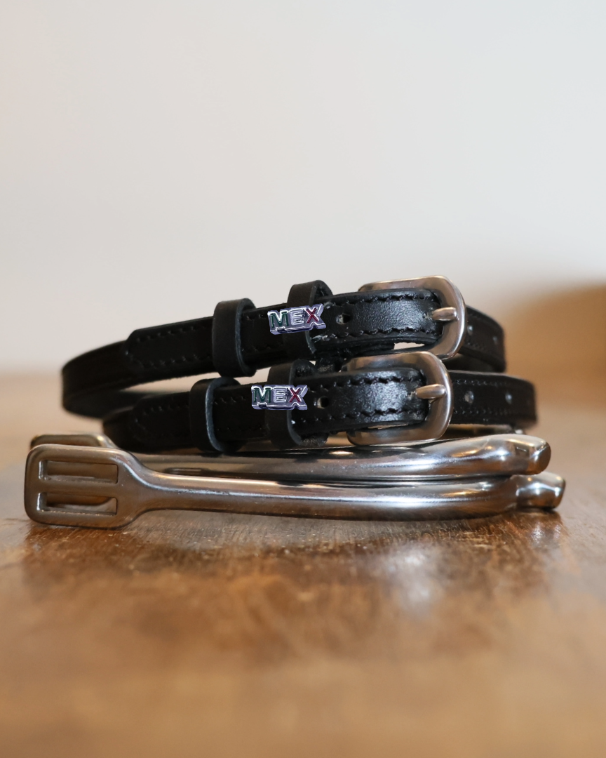 Black leather MEXico spur straps coiled beside a silver spur on a wooden surface