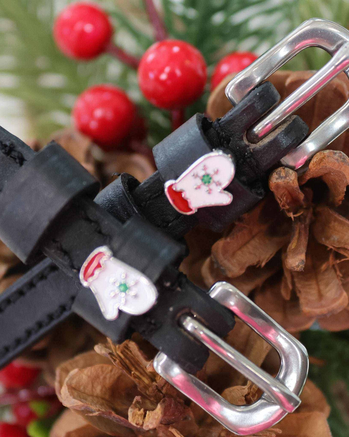 Close-up of white winter mitten charms on black leather spur straps among pine cones and berries