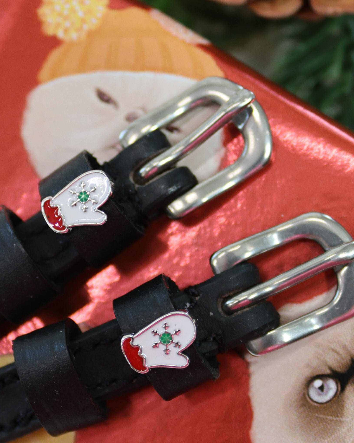 Angled close-up of white mitten charms on black leather spur straps against a red holiday box