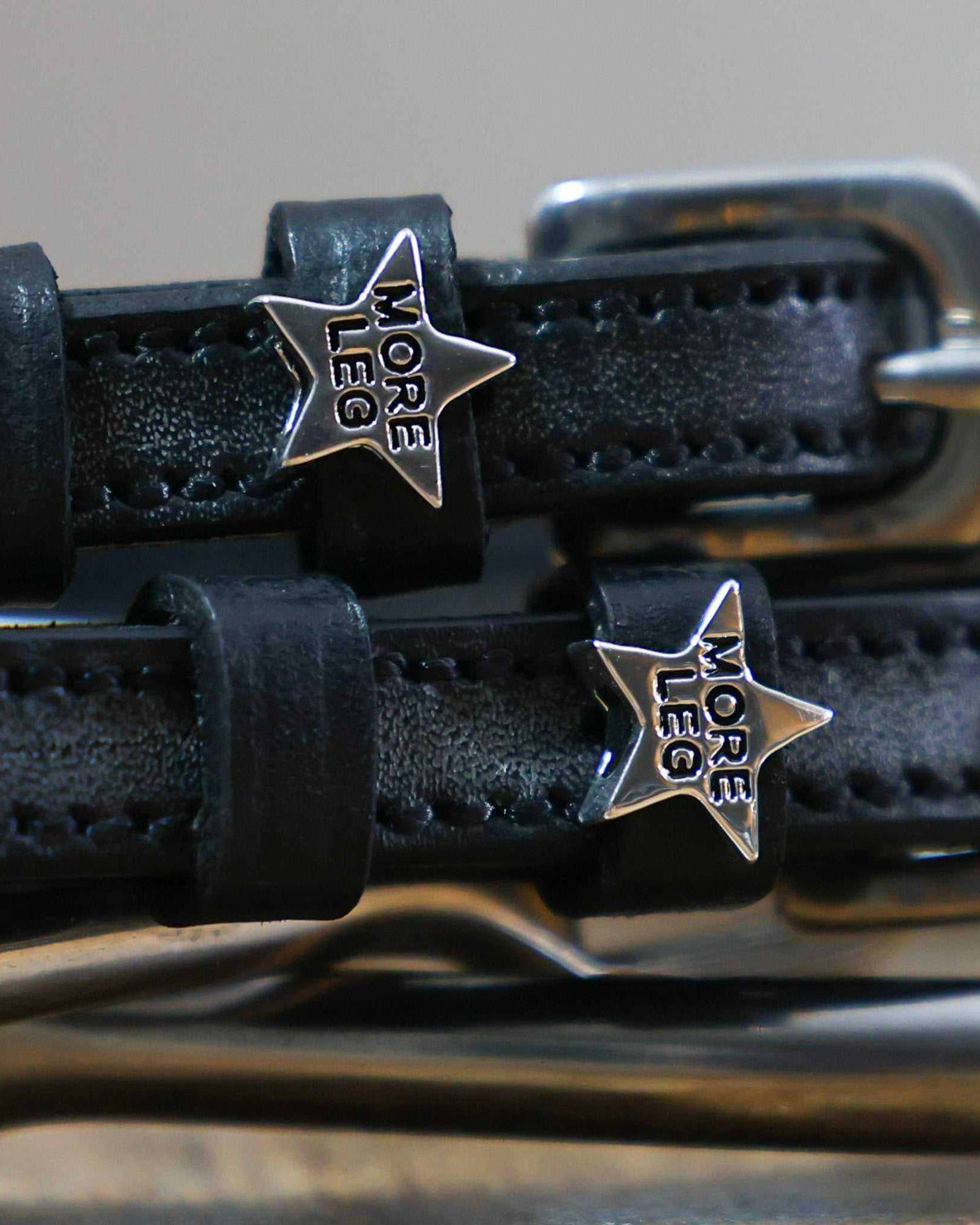 Close-up of silver MORE LEG star charms on black leather spur strap keepers