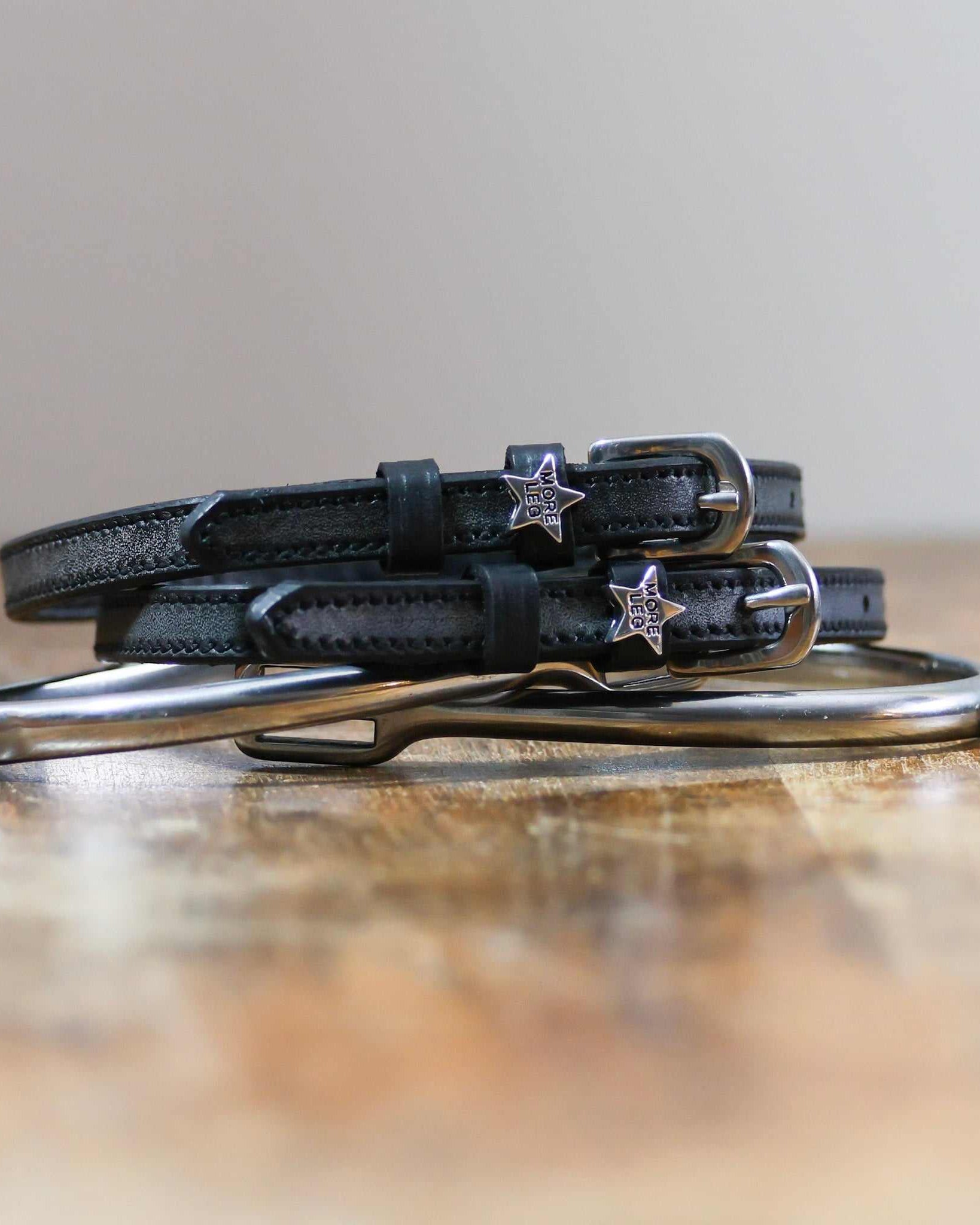 Black MORE LEG star spur straps stacked on a silver spur on wood, close-up