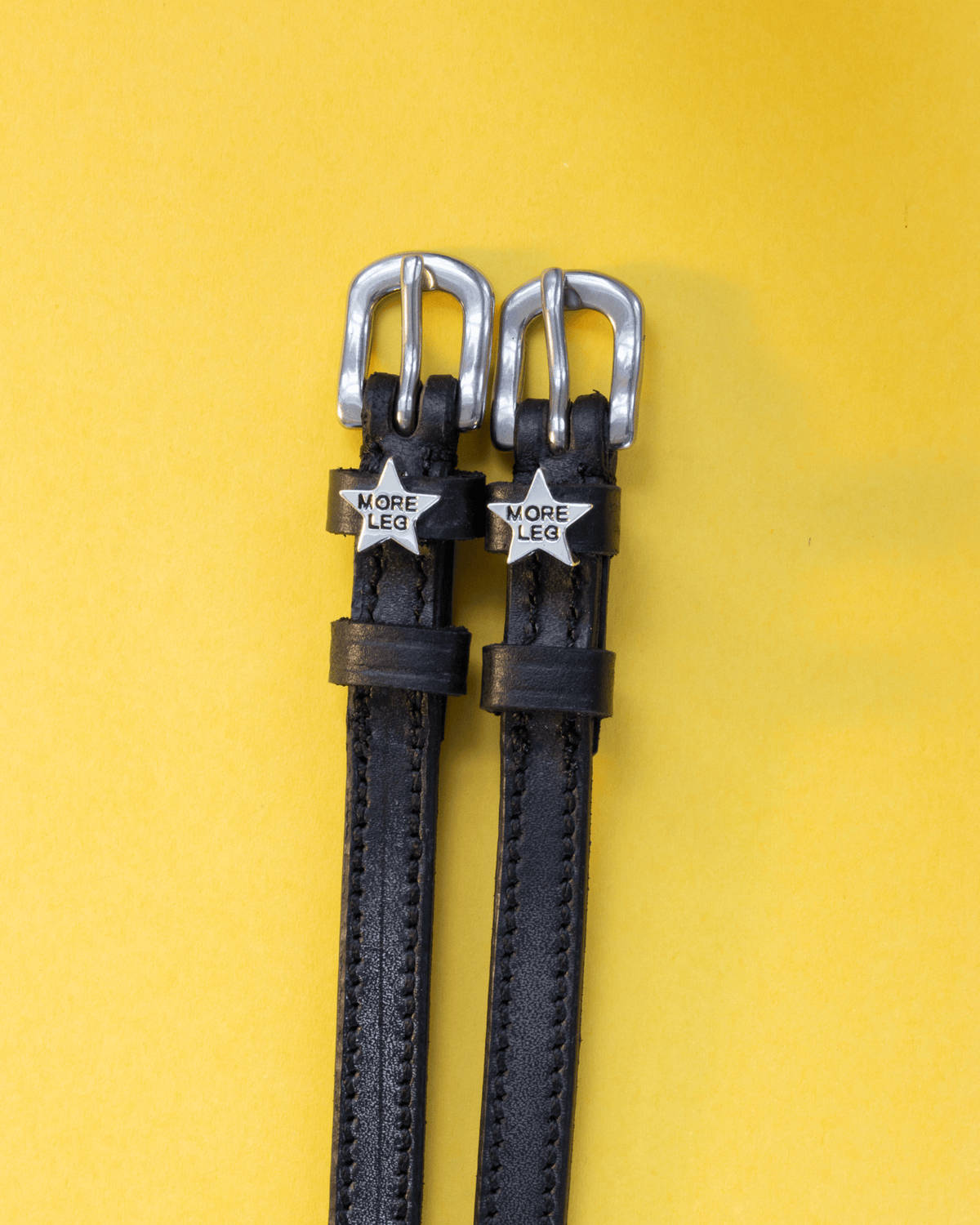 Pair of black leather MORE LEG star spur straps flat-laid on yellow