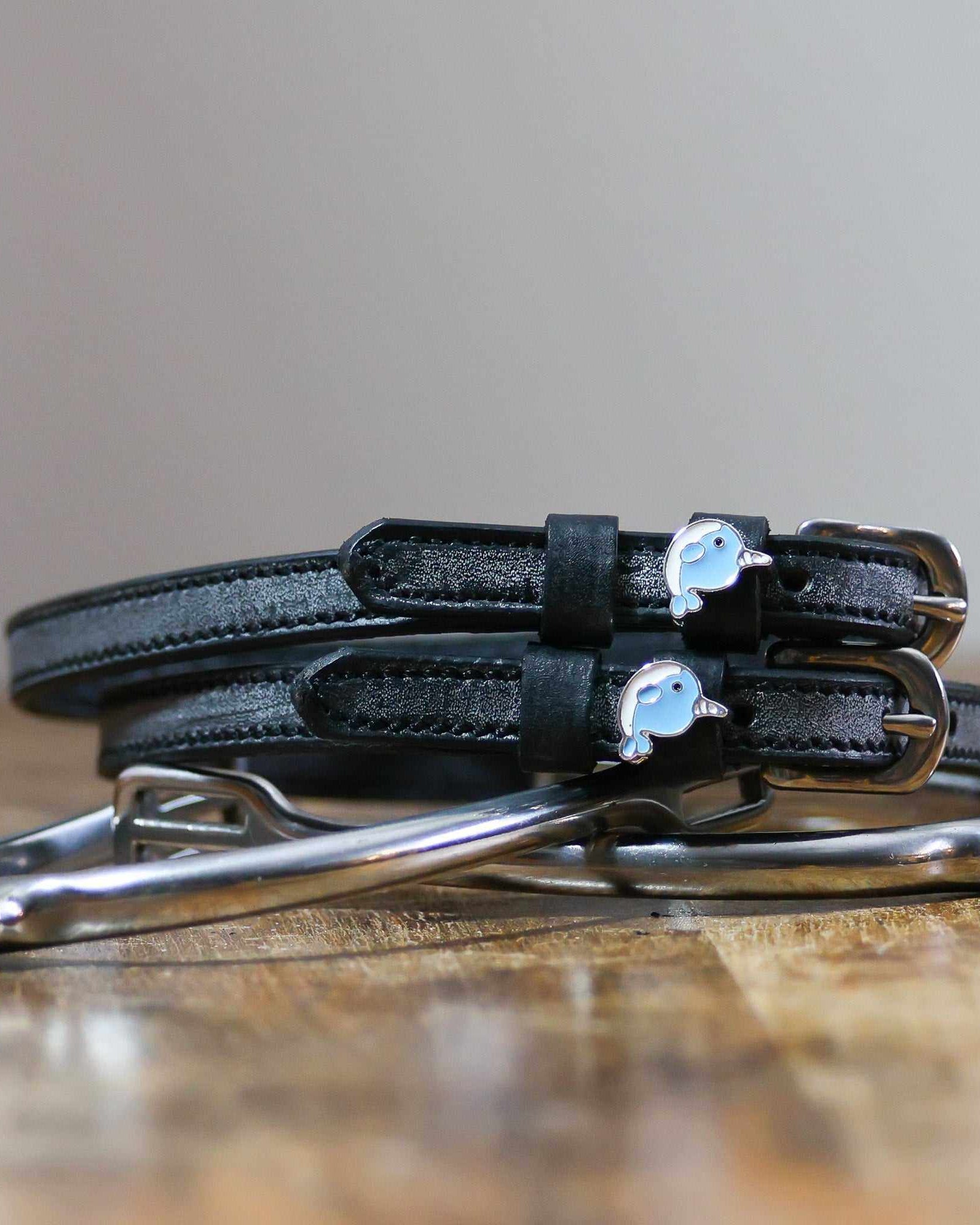 Black leather narwhal spur straps coiled on a wooden surface beside a silver bit