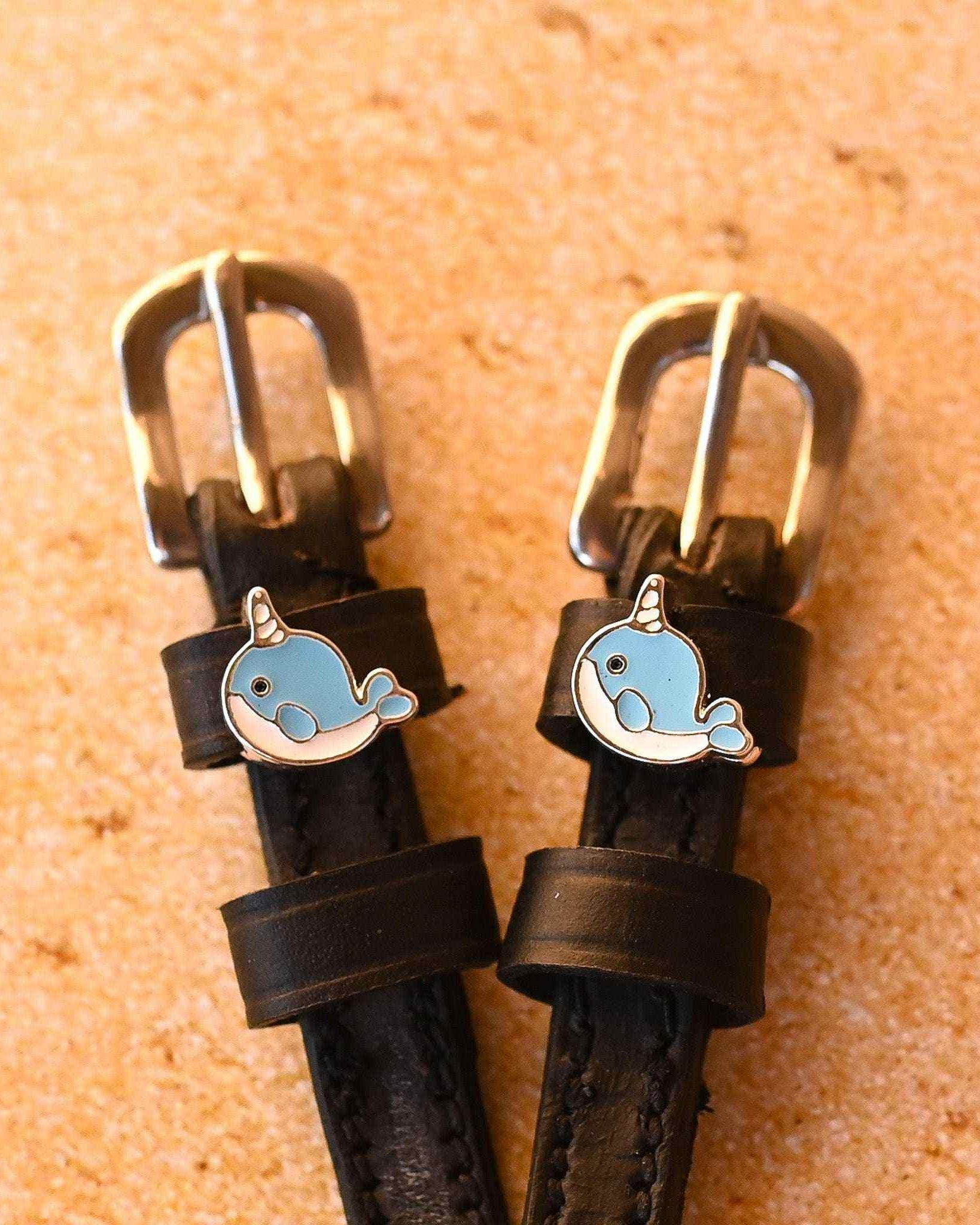 Black leather narwhal spur straps with brass buckles on a sandy cork background