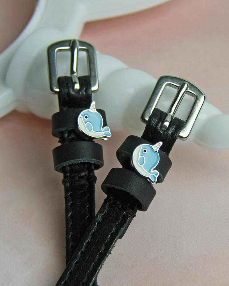 Narwhal charm spur straps draped over a white stand on a pink background