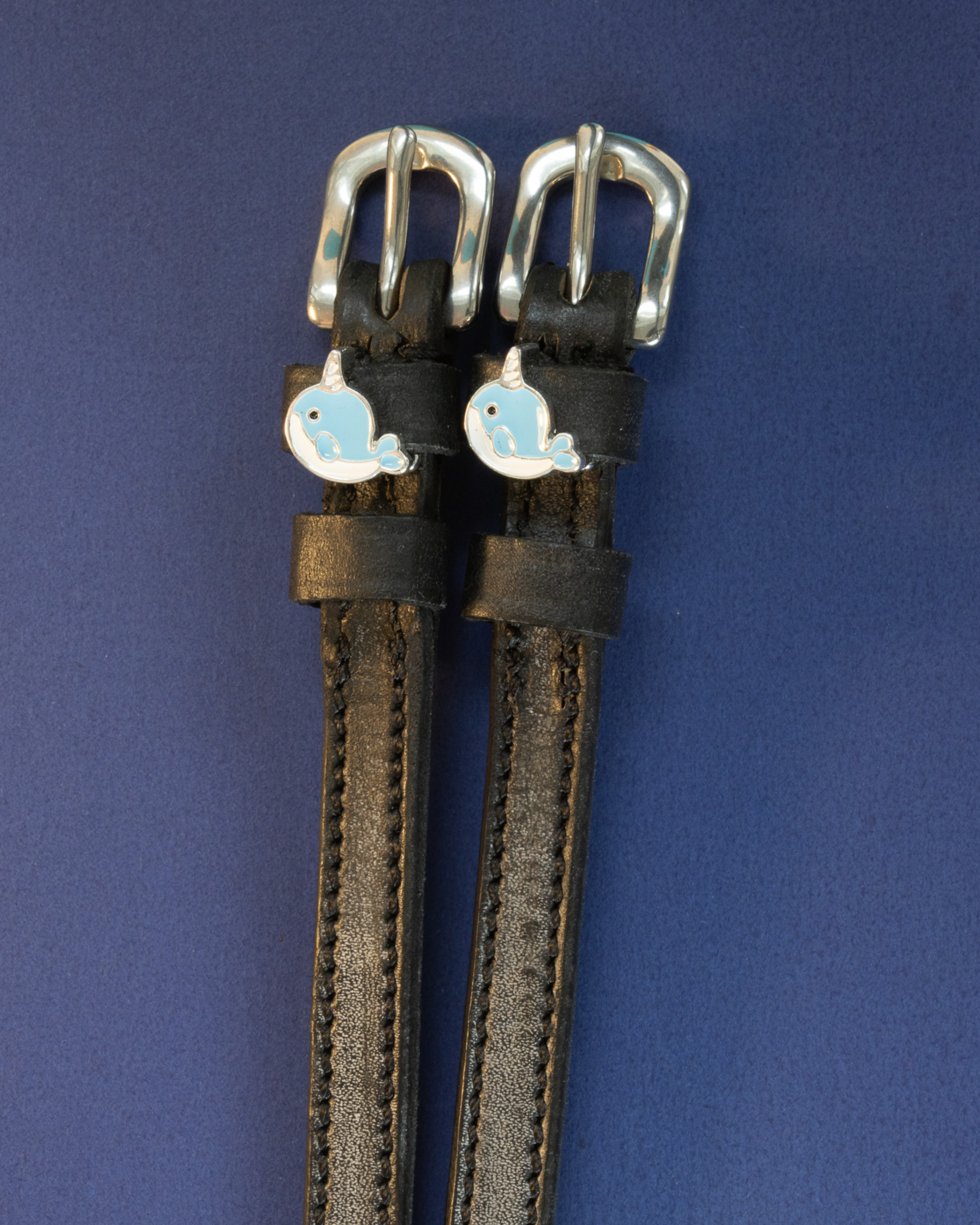 Black leather narwhal charm spur straps flat-laid on a navy blue background