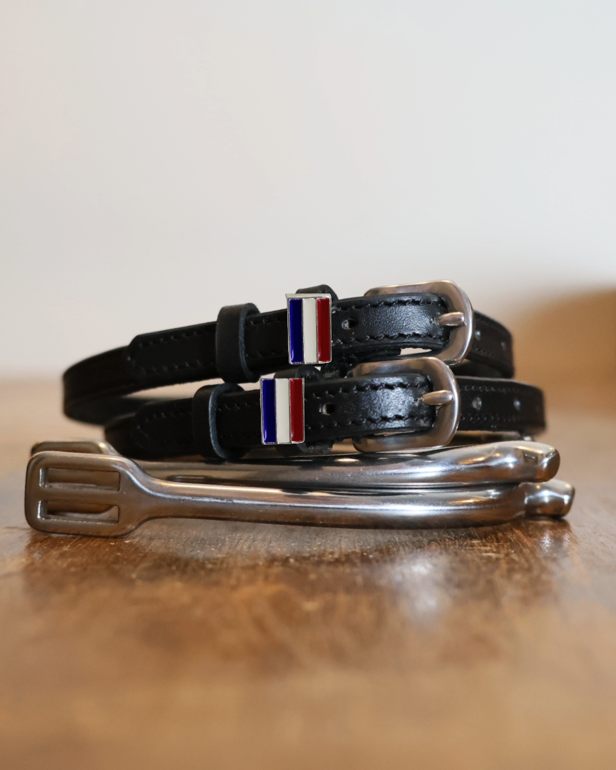 Black leather Netherlands flag spur straps coiled beside a stirrup on a wooden surface