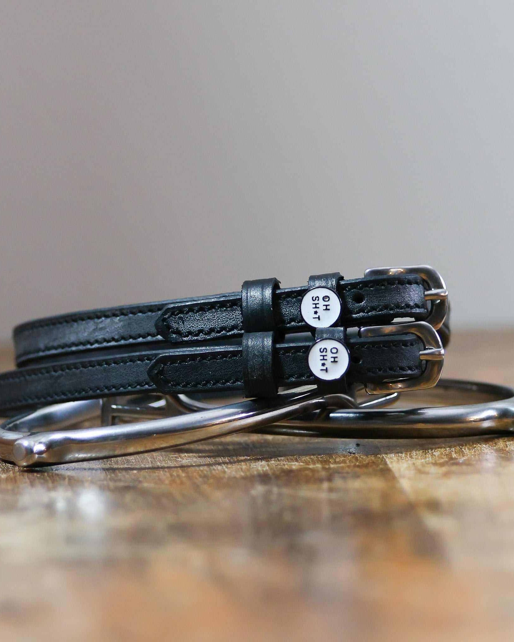 Black leather OH SH*T spur straps coiled beside a stirrup on wood