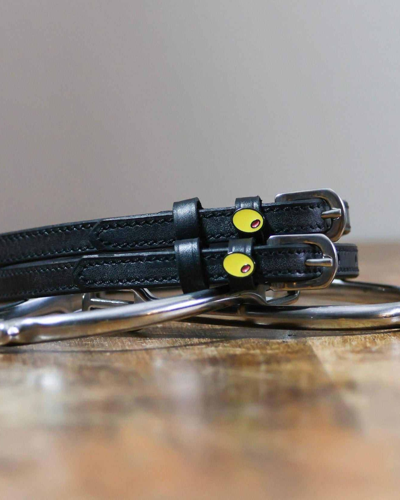 Black leather olive spur straps coiled on a wooden surface with a silver bit