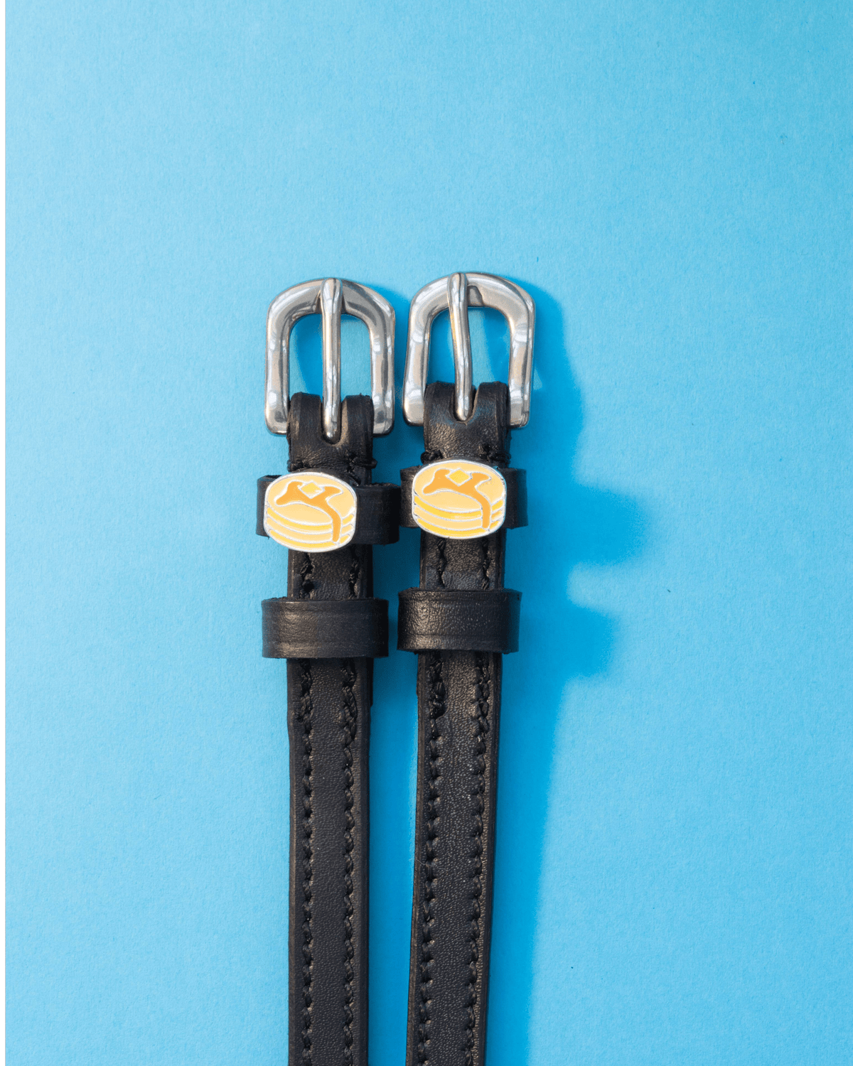 Black leather pancake spur straps flat-laid on a bright blue background