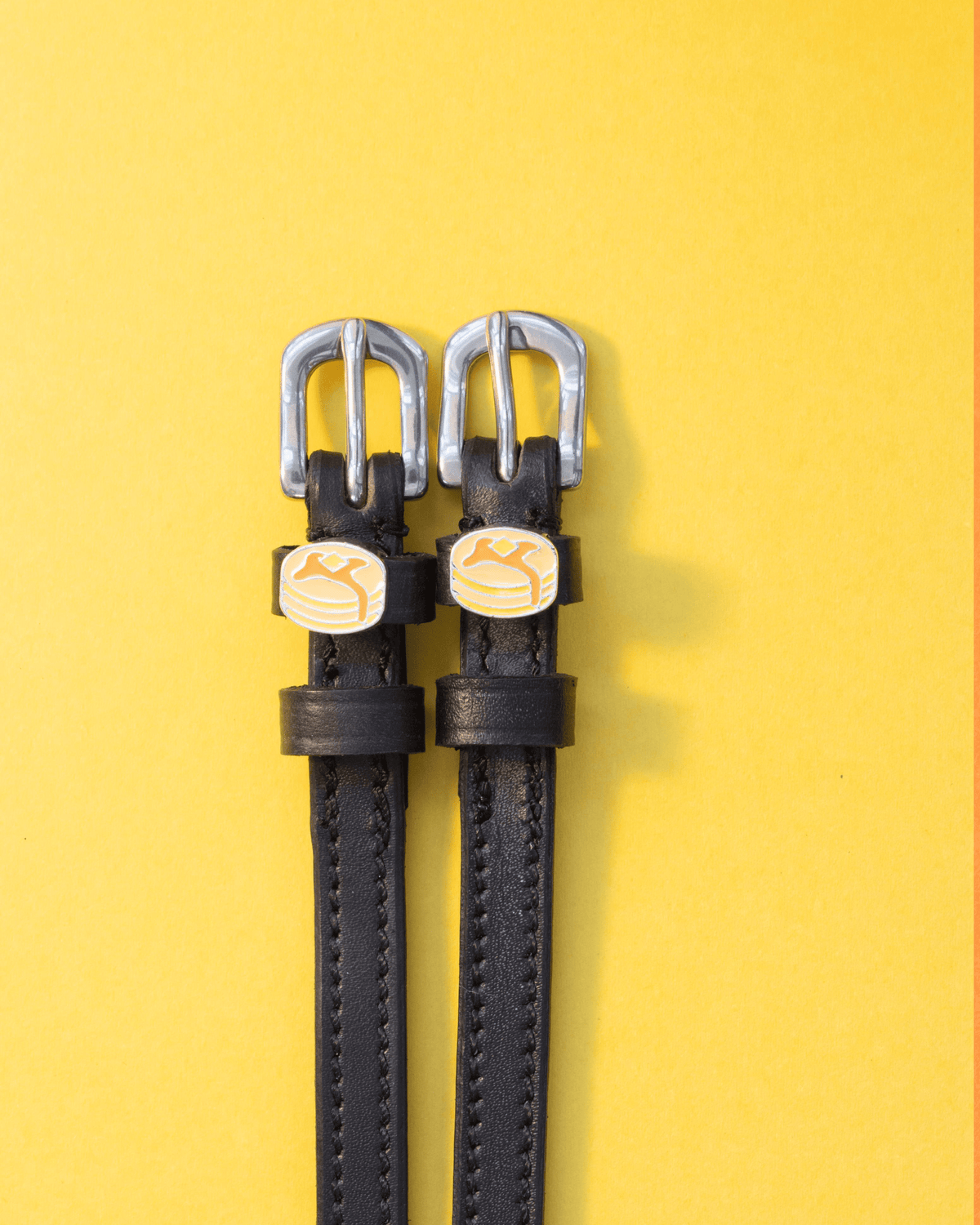 Pair of black leather ManeJane pancake spur straps on a yellow background