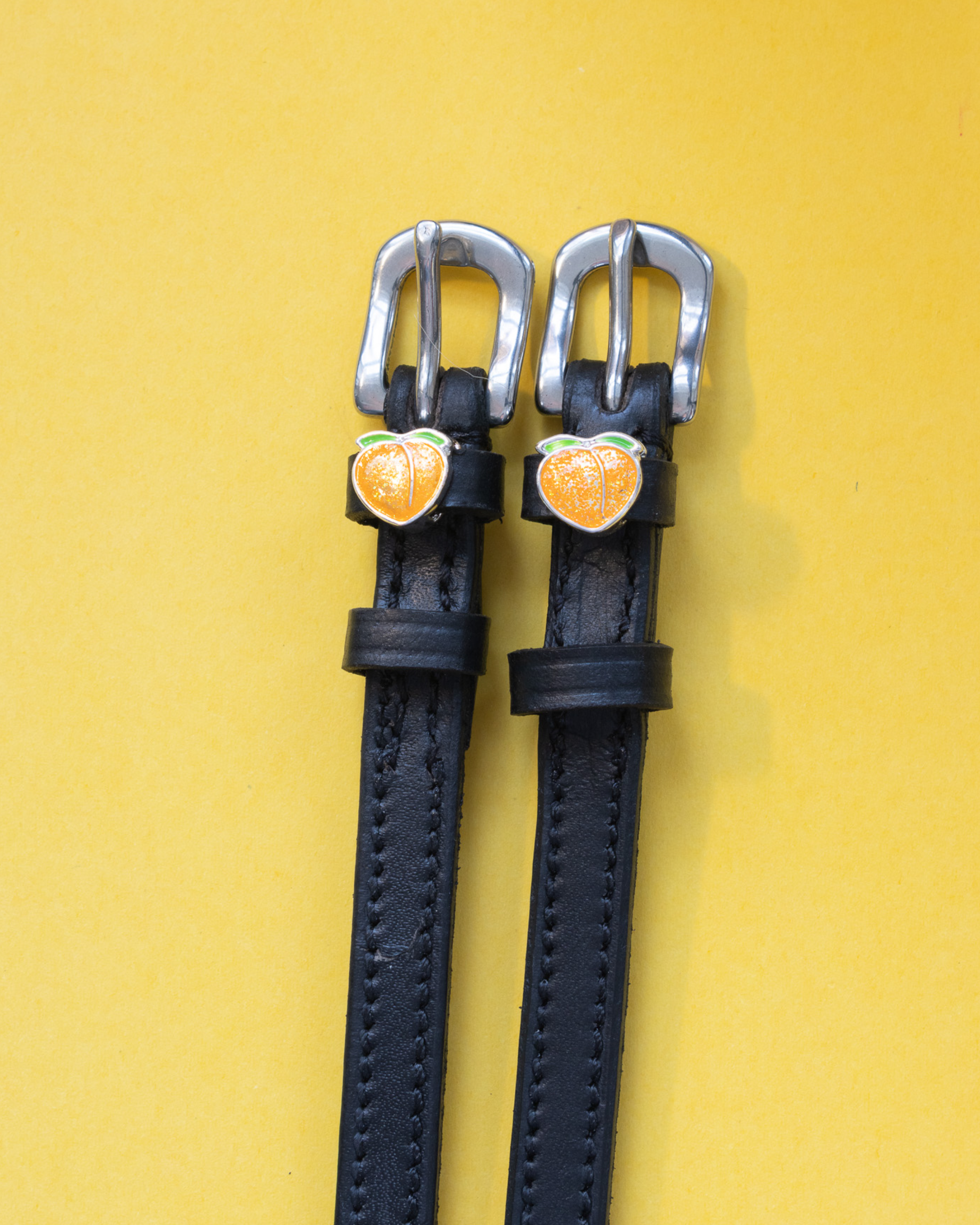 Black leather peach charm spur straps flat-laid on a yellow background