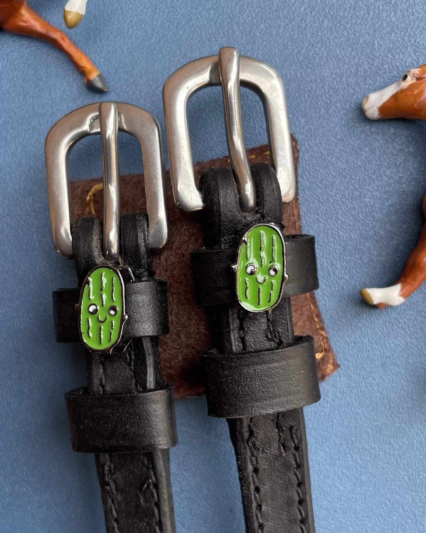 Green pickle charm spur straps with toy horses on blue, ManeJane lifestyle flat-lay
