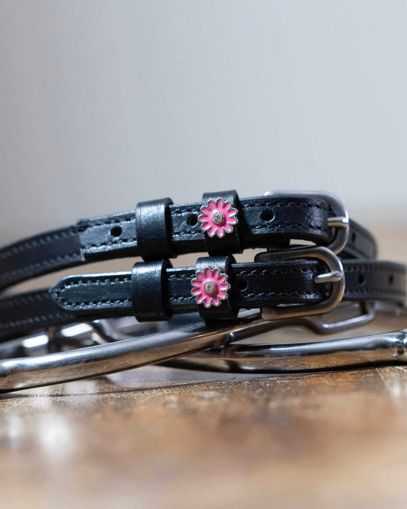 Pink flower charm spur straps coiled beside a stirrup on a wooden tabletop