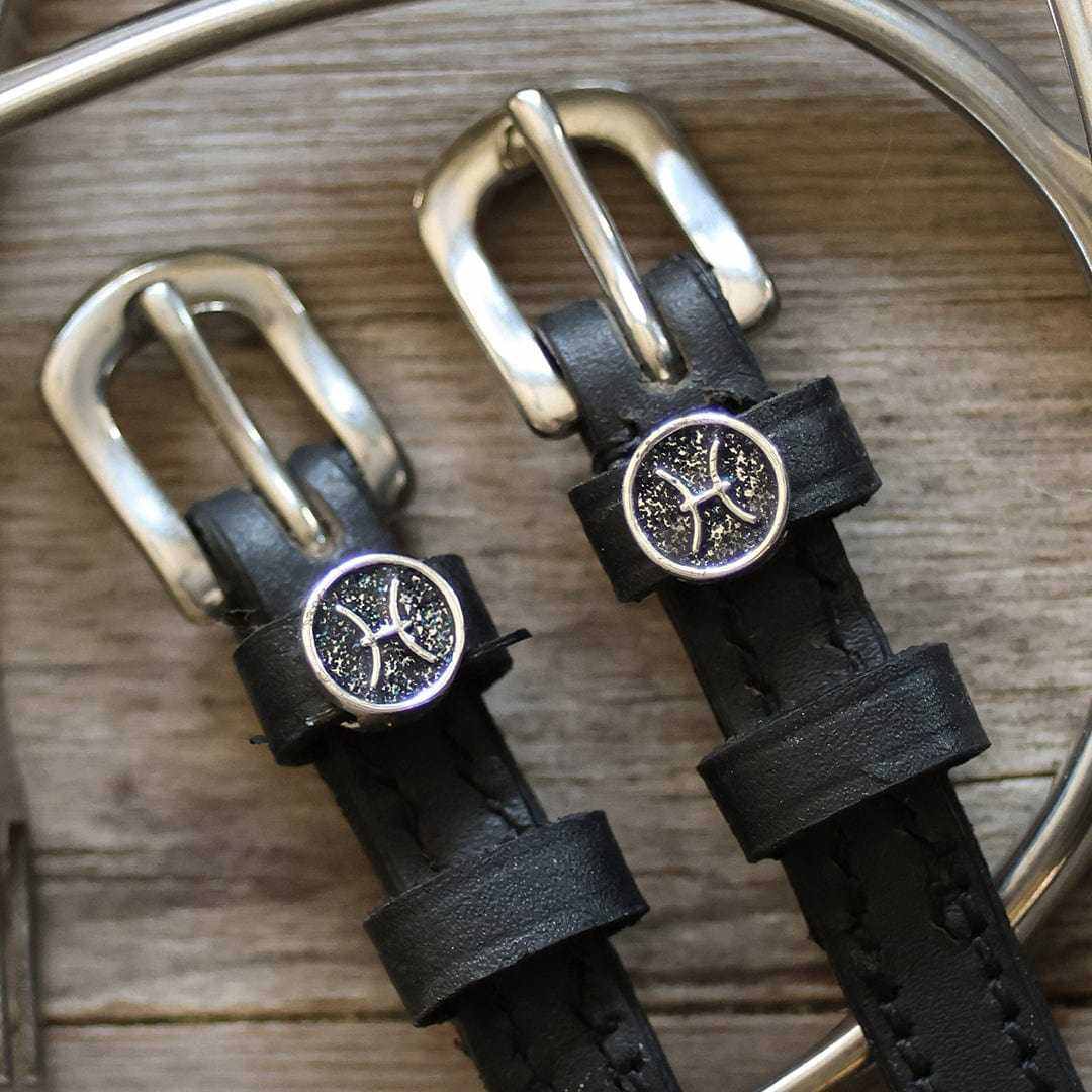 Close-up of Pisces zodiac charms and silver buckles on black spur straps, wood