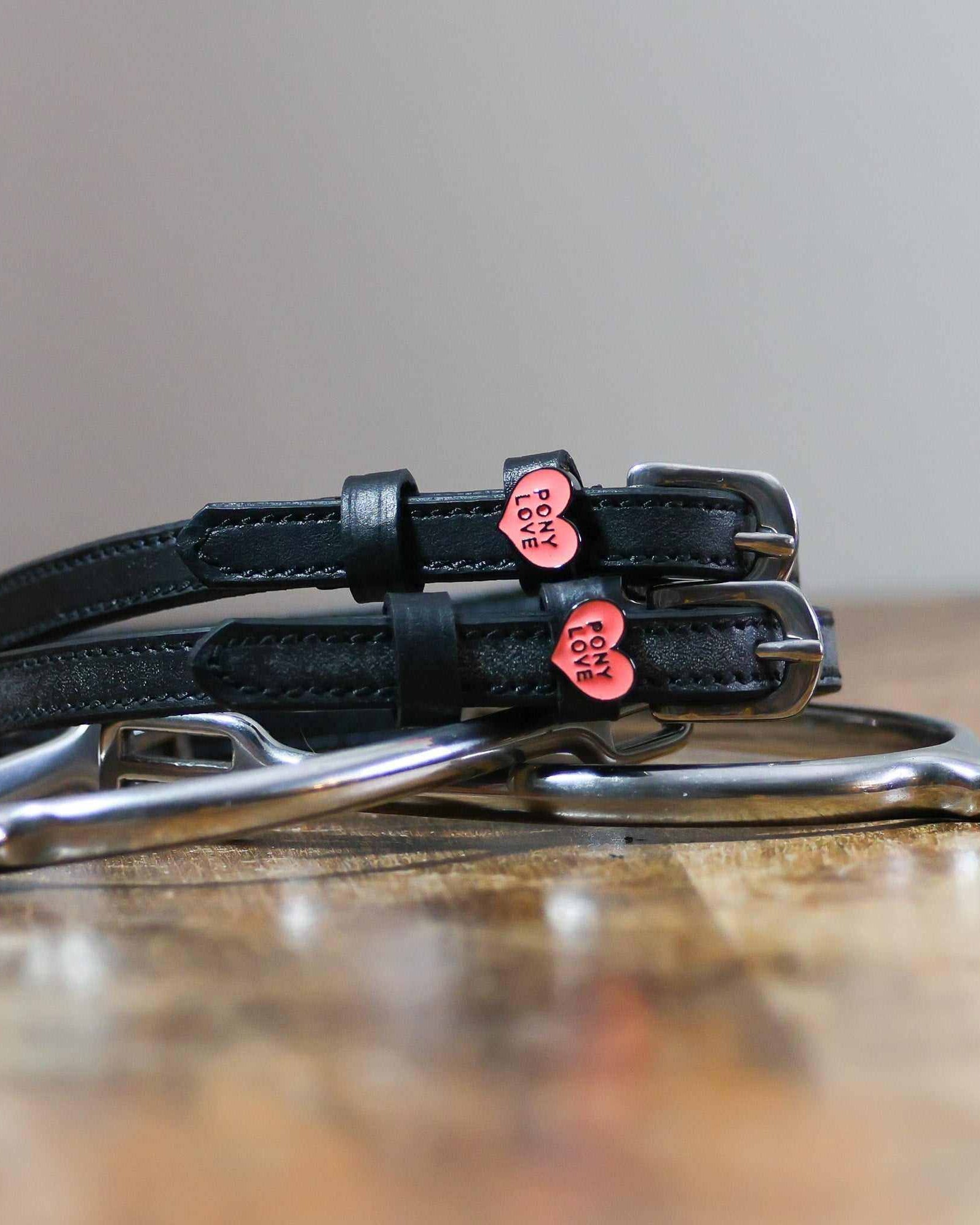 Pony Love heart-charm spur straps coiled beside a stirrup on a wooden tabletop