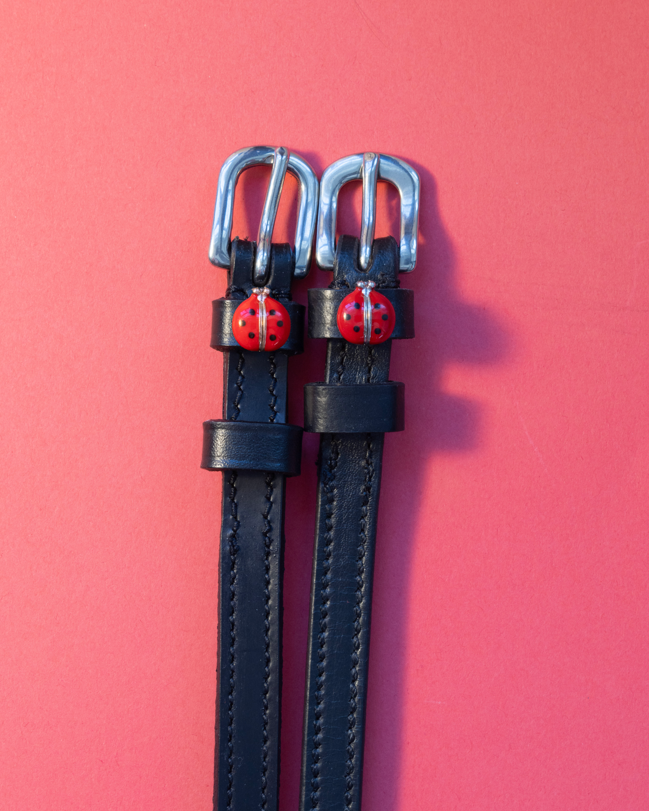 Pair of red ladybug charm spur straps laid flat on a coral background