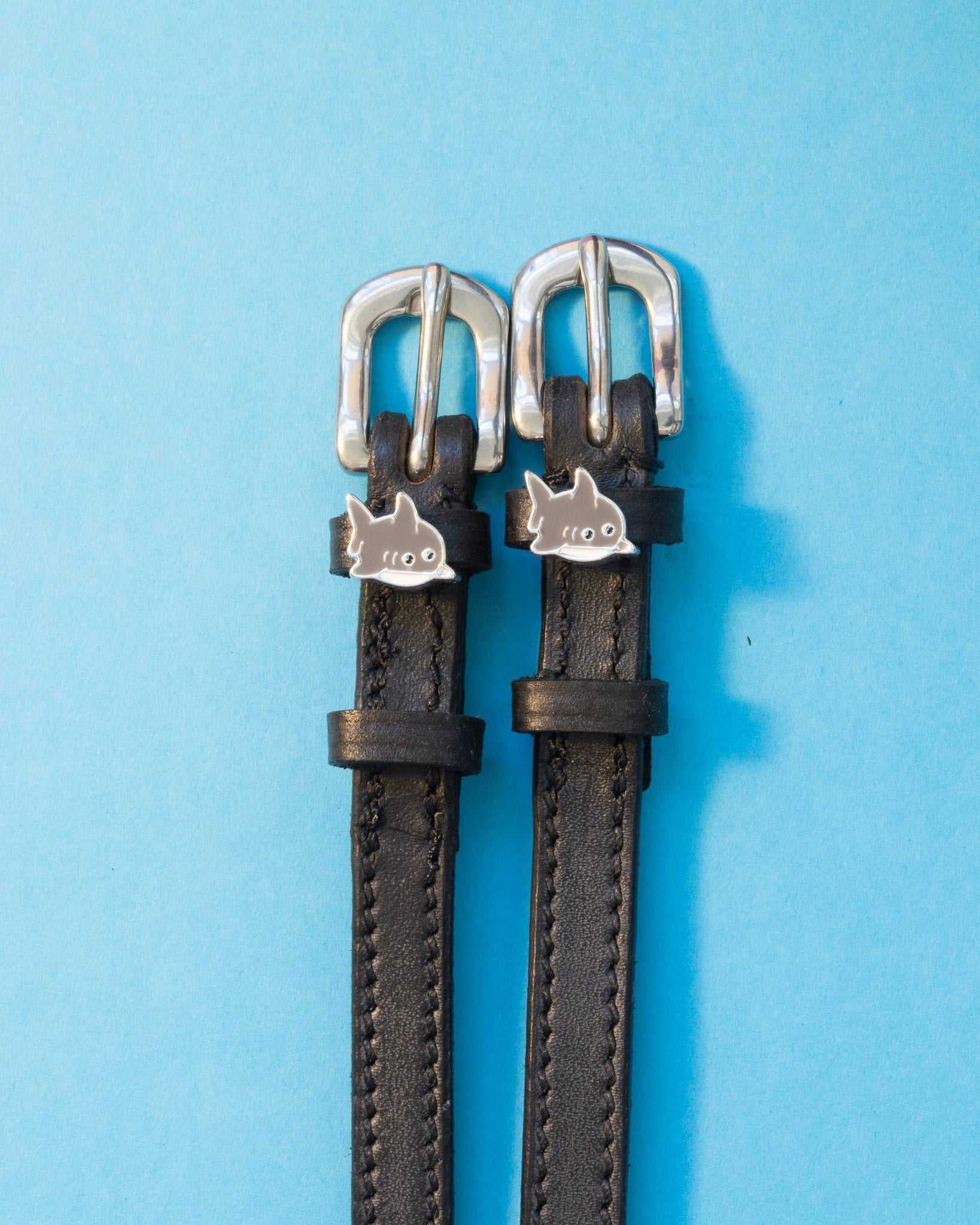 Black leather shark charm spur straps with silver buckles on a light blue background