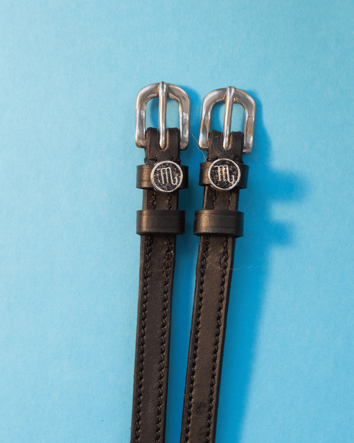 Black leather ManeJane spur straps with silver buckles and round Scorpio zodiac charms on blue