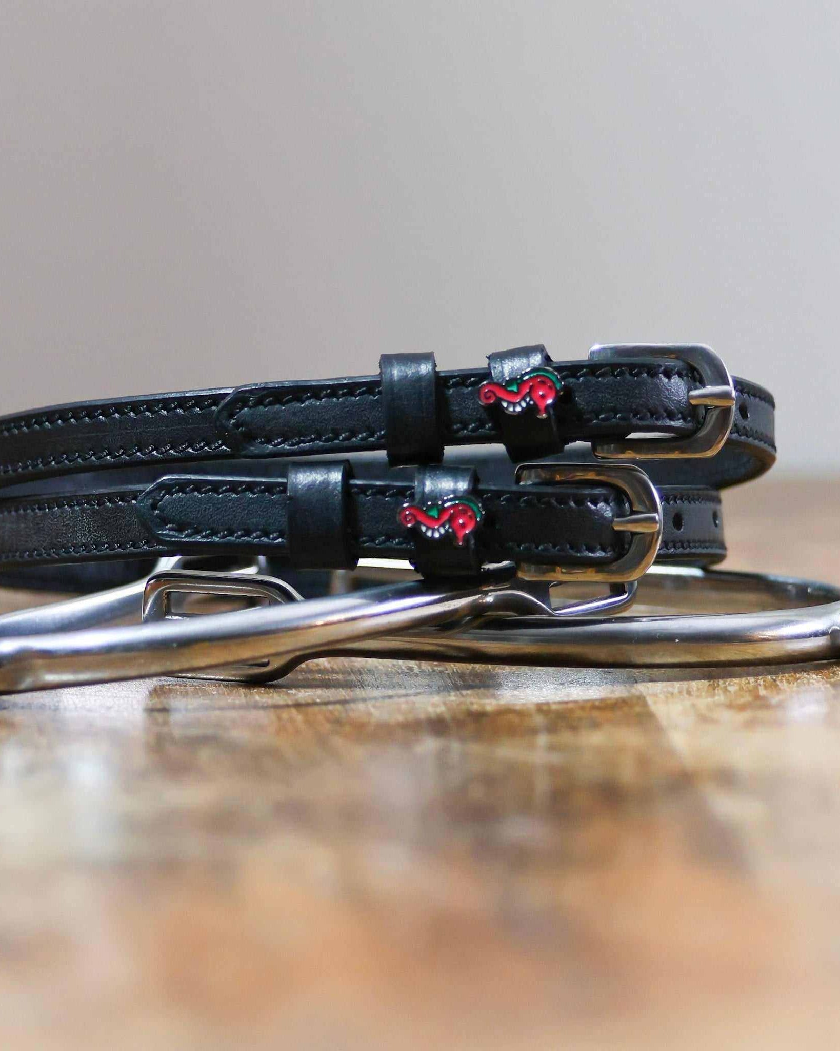 Black leather seahorse spur straps coiled beside a stirrup on a wooden surface