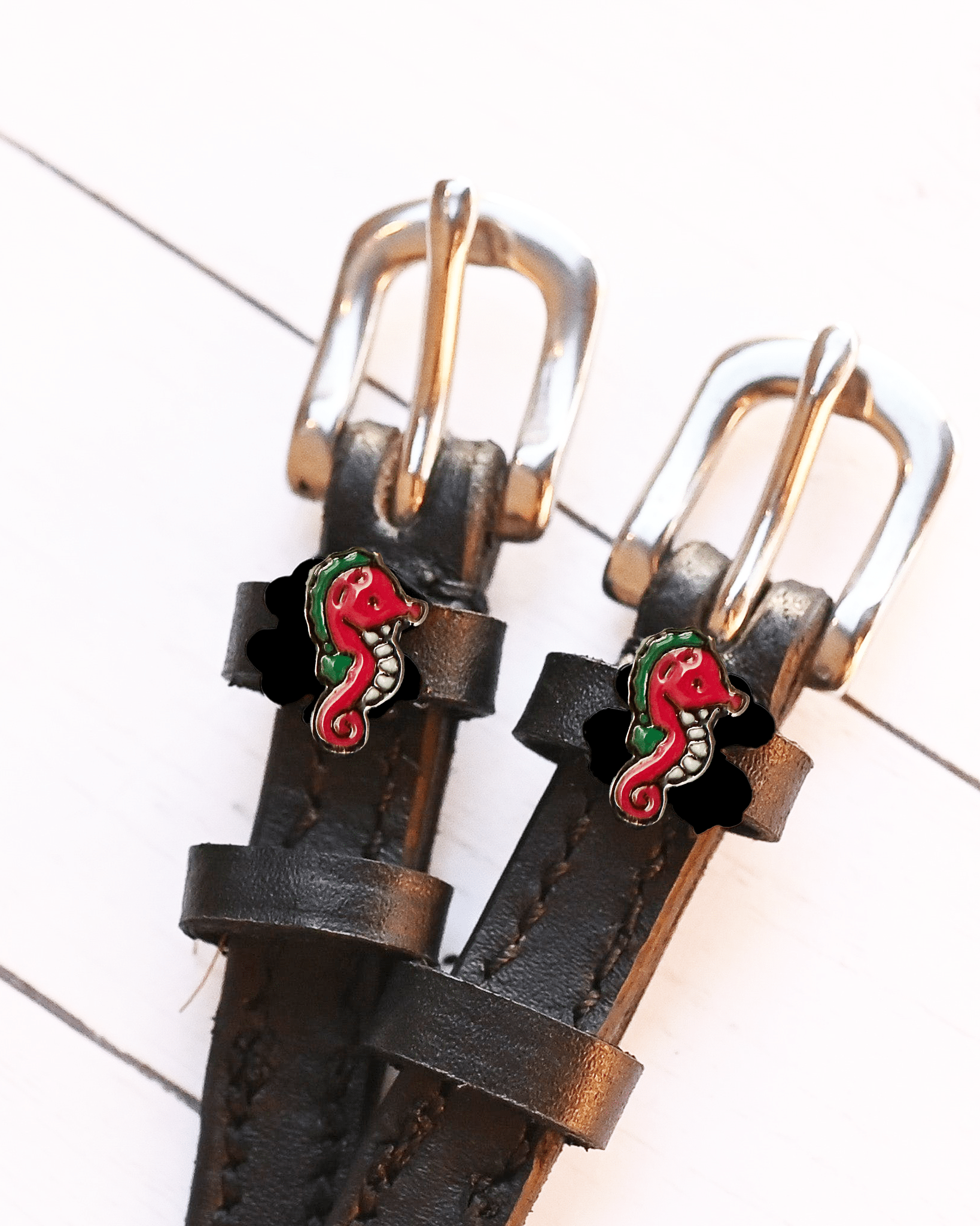 Angled close-up of red seahorse charms on ManeJane spur straps over white planks