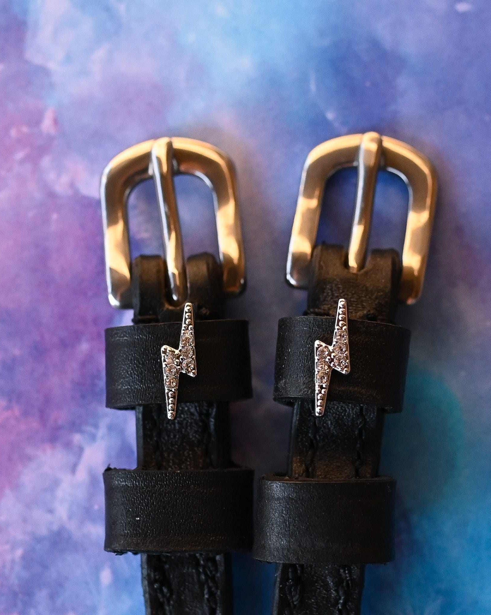 Close-up of rhinestone lightning bolt charms on black spur straps over a galaxy backdrop