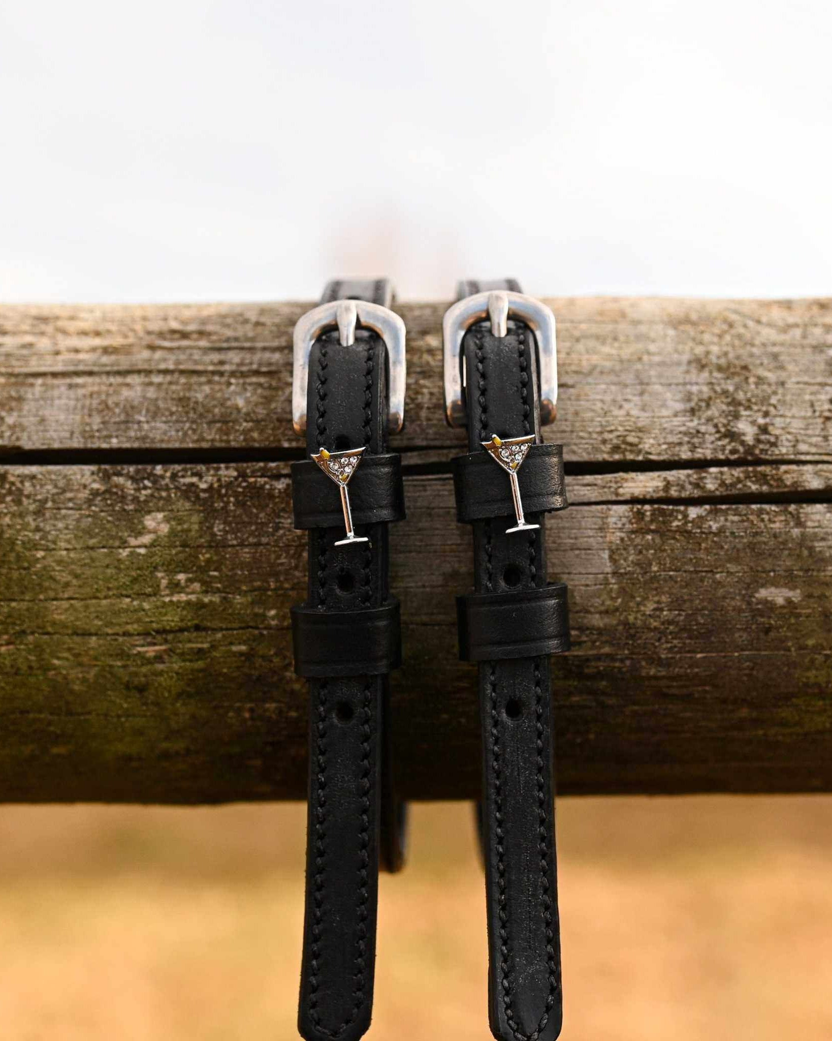 ManeJane martini glass spur straps hanging over a weathered wooden fence rail