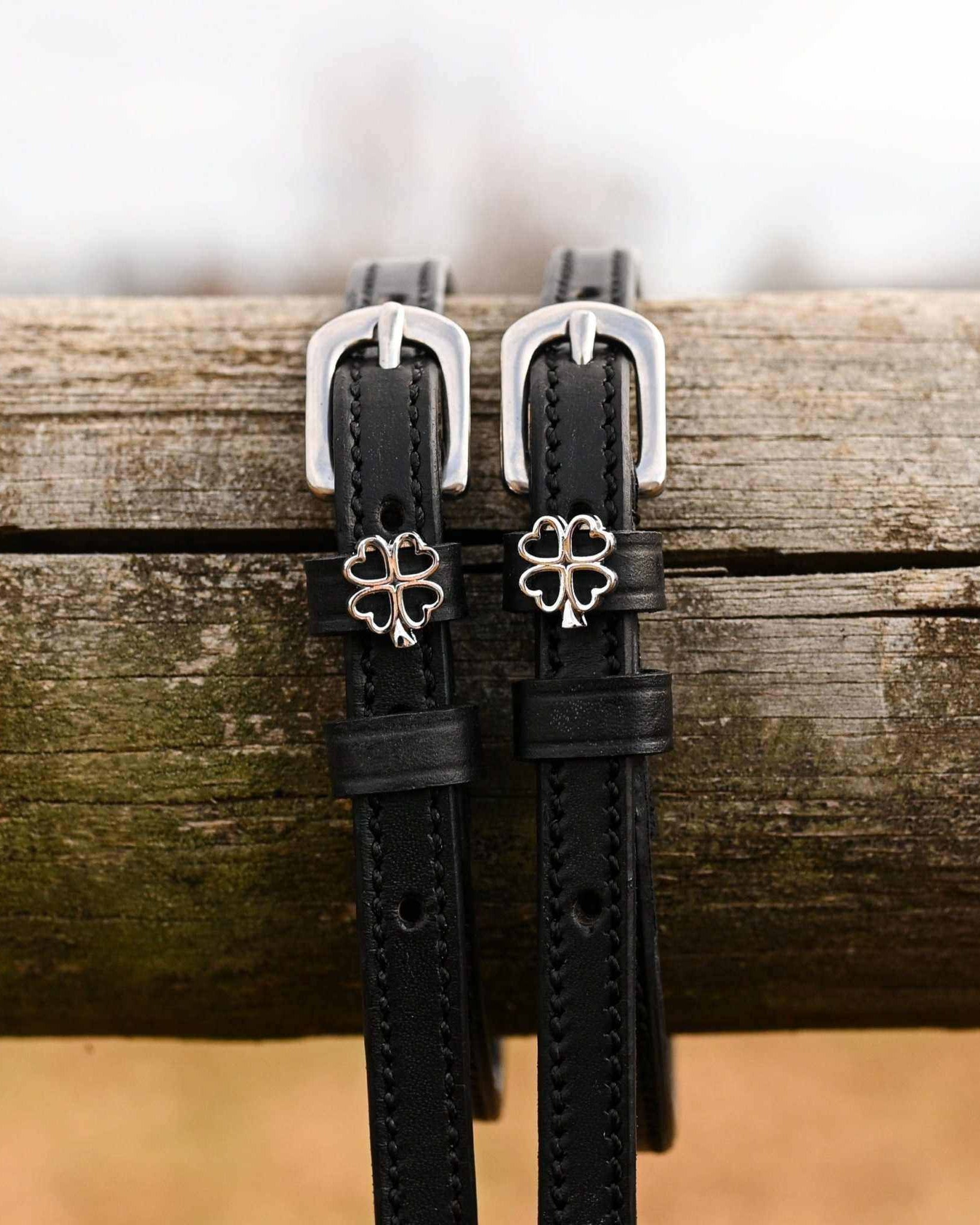 Silver outline clover charms on black leather spur straps draped over a wooden fence rail