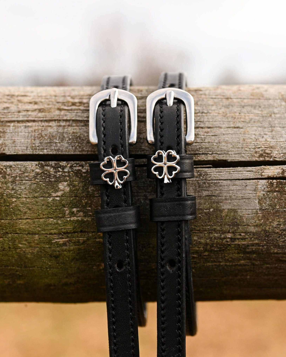 Silver outline clover charms on black leather spur straps draped over a wooden fence rail
