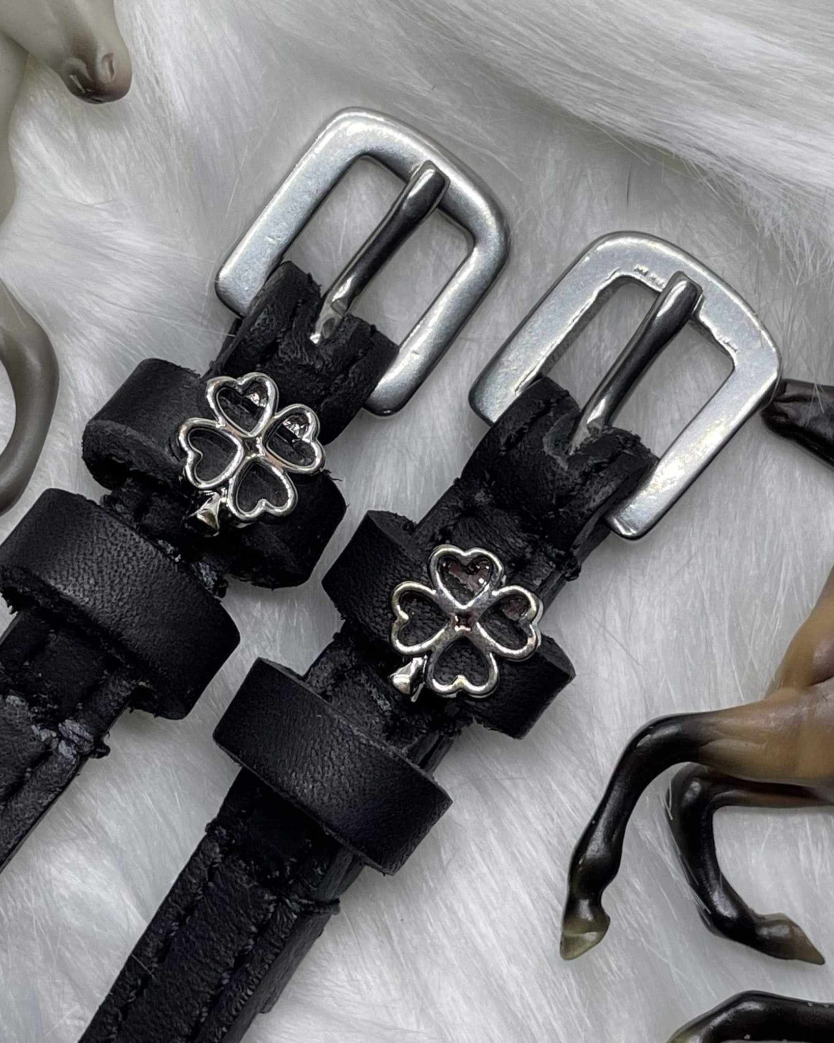 Black leather silver clover spur straps on white fur beside toy horse figurines