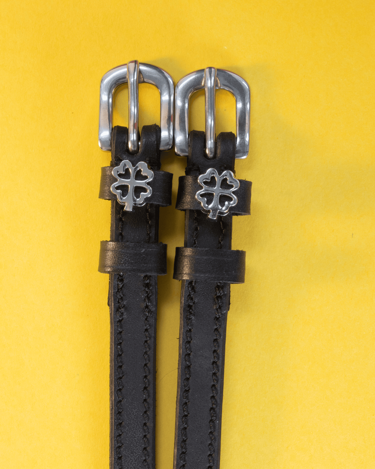 Silver openwork clover charms on black leather ManeJane spur straps, yellow background
