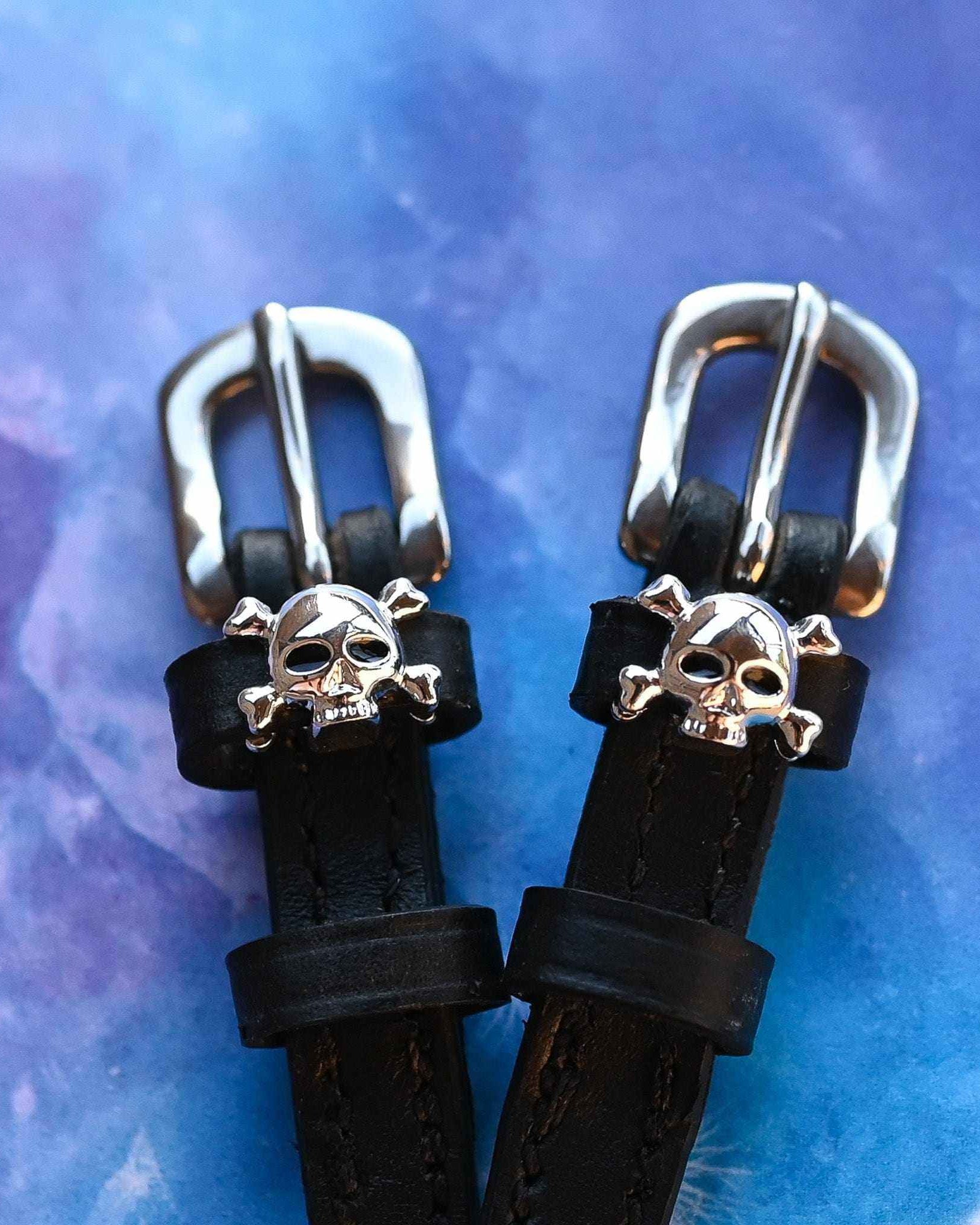 Close-up of silver skull and crossbones charms on spur straps over a purple marble backdrop