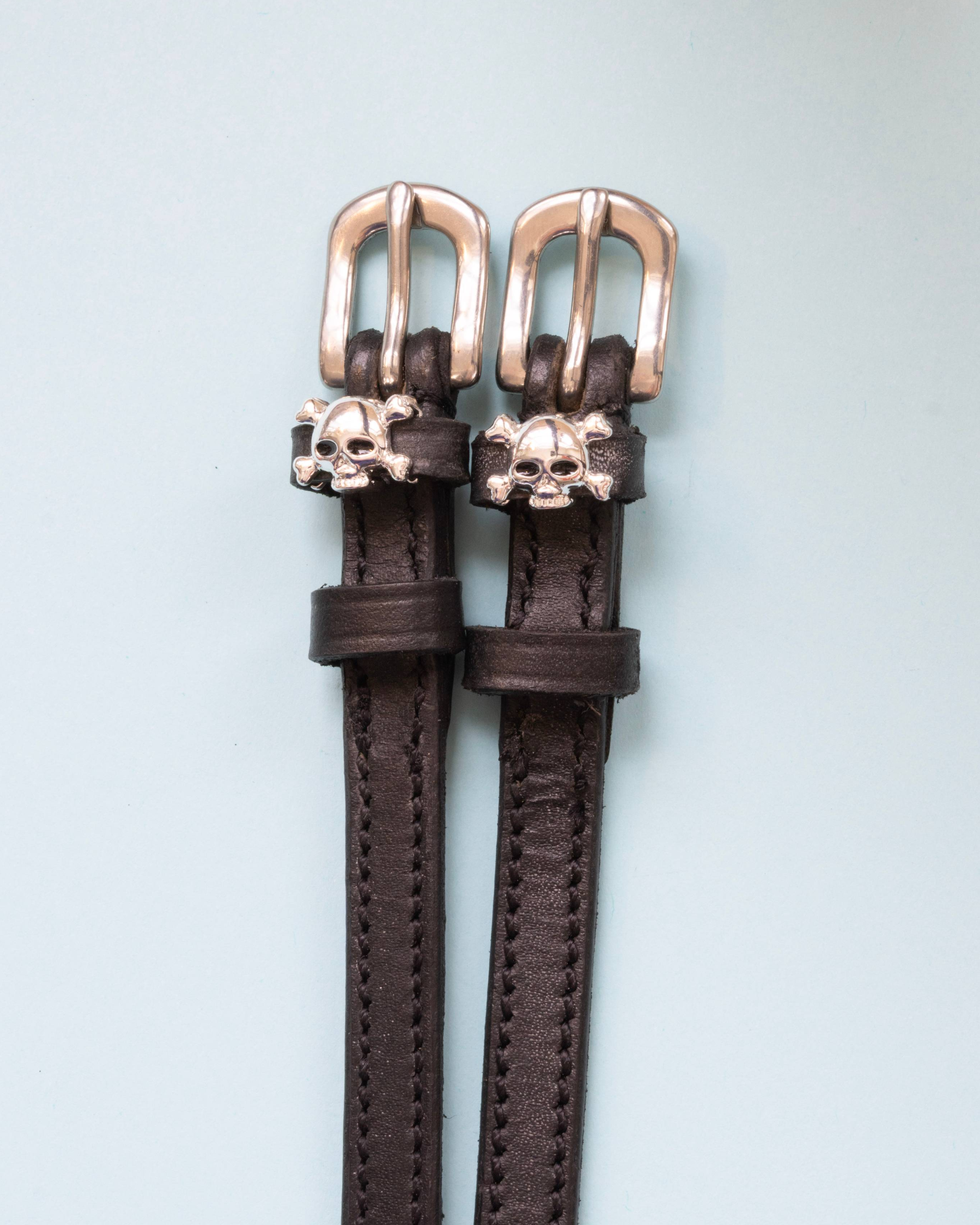 Brown leather skull and crossbones spur straps with rose gold buckles on pale blue