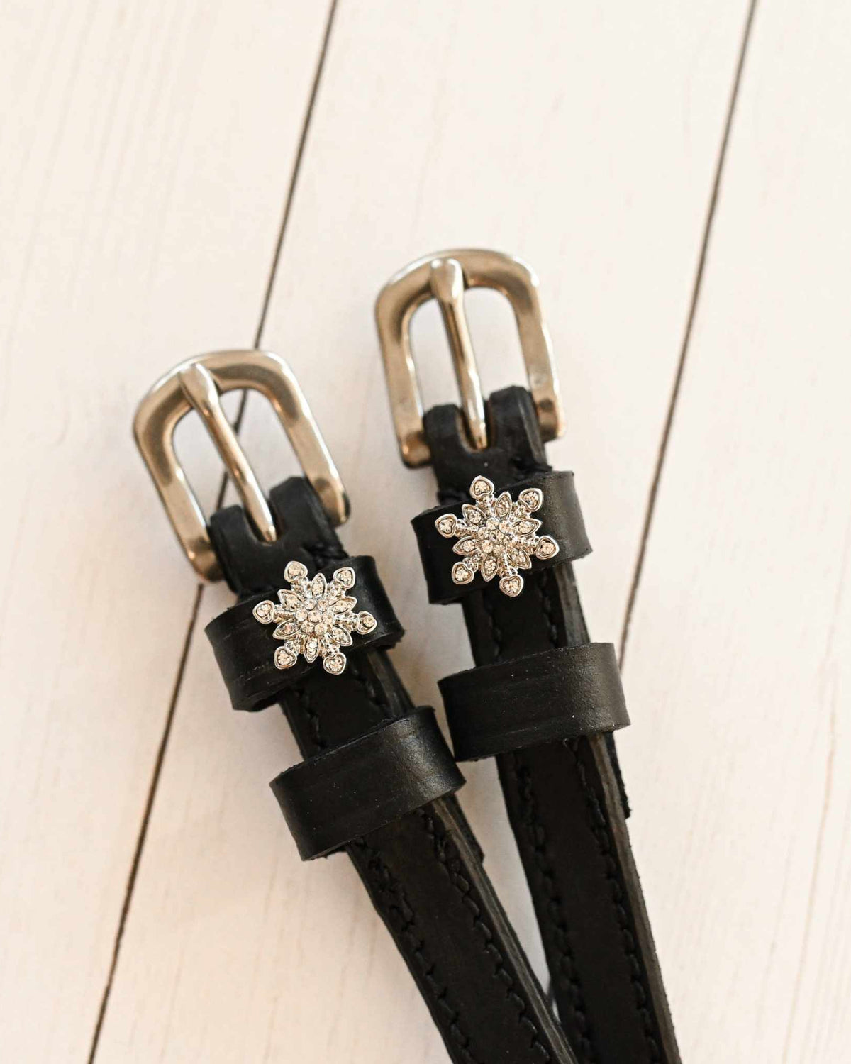 Close-up of black snowflake spur straps with crystal charms on white-washed wood planks