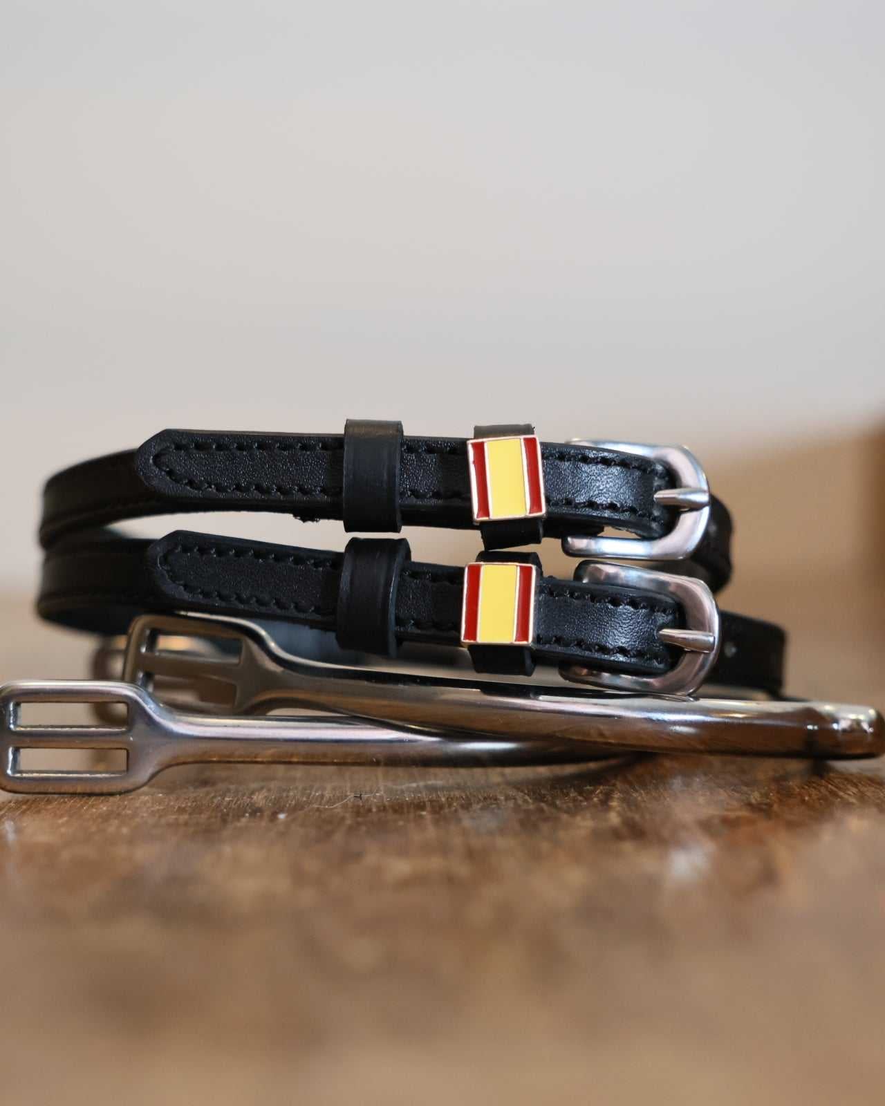 Spanish flag charm spur straps coiled beside a stirrup on a wooden tabletop