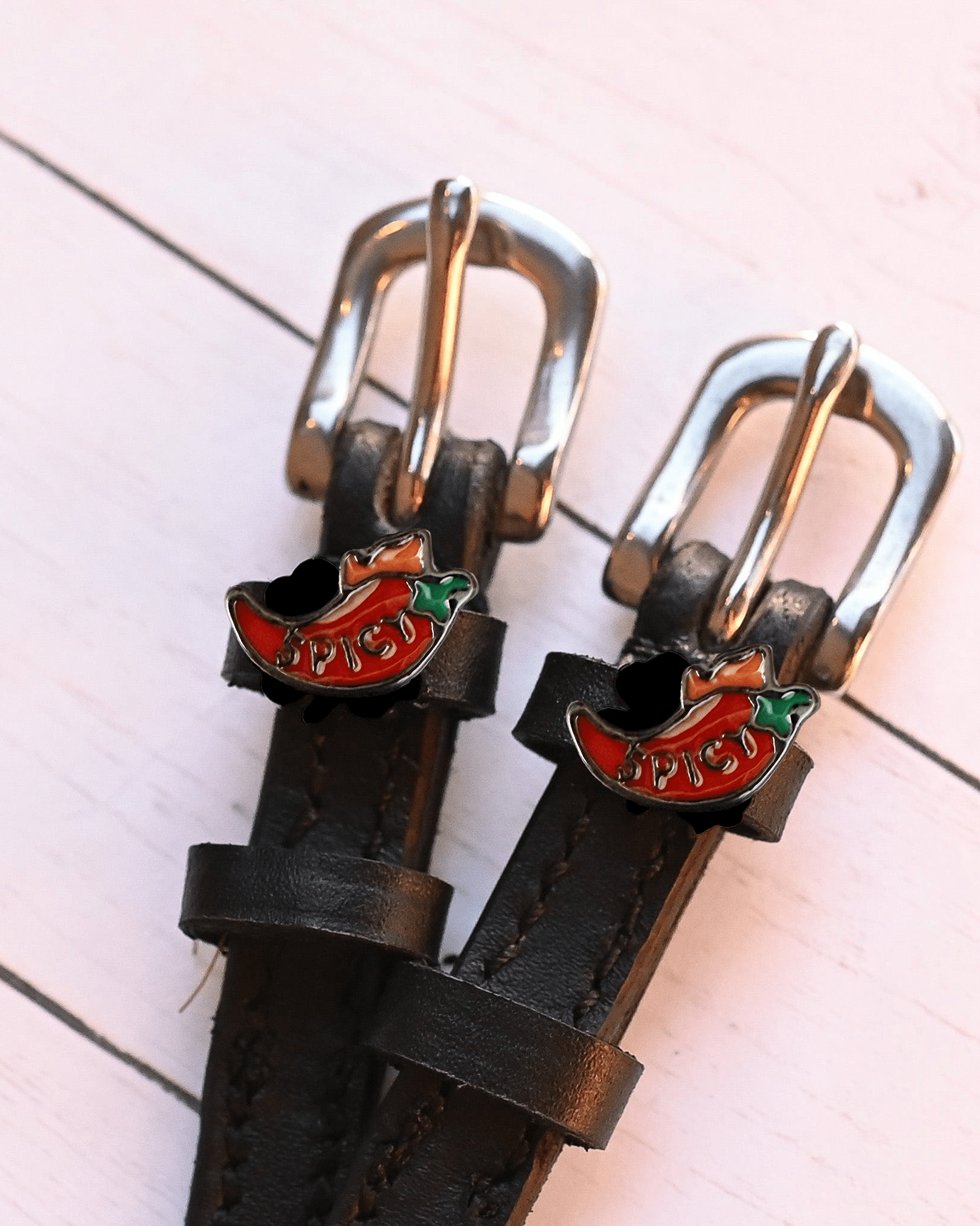 Angled close-up of red 'SPICY' chili charms on black leather ManeJane spur straps