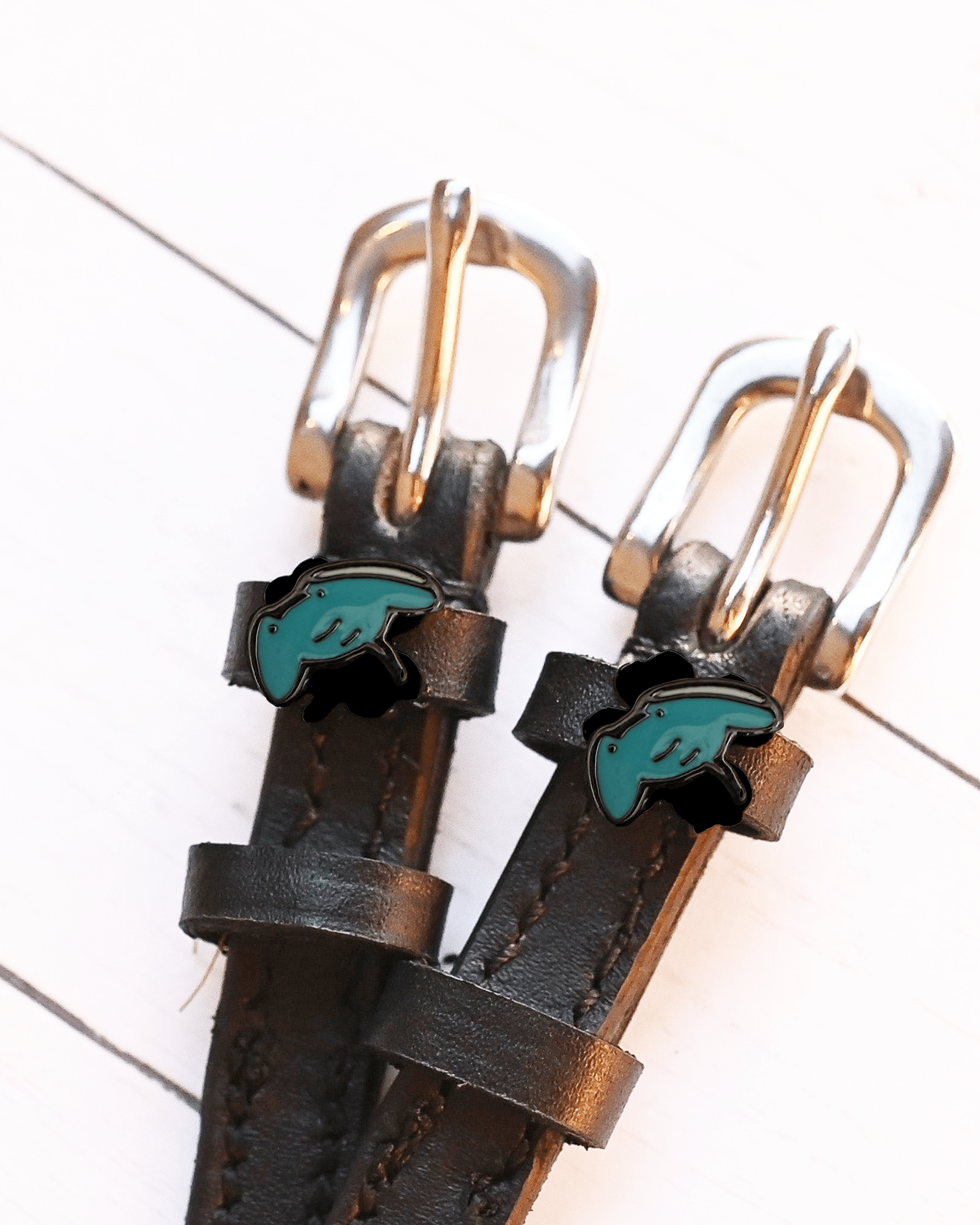 Teal stingray charms on black leather spur straps laid on white wood planks, ManeJane