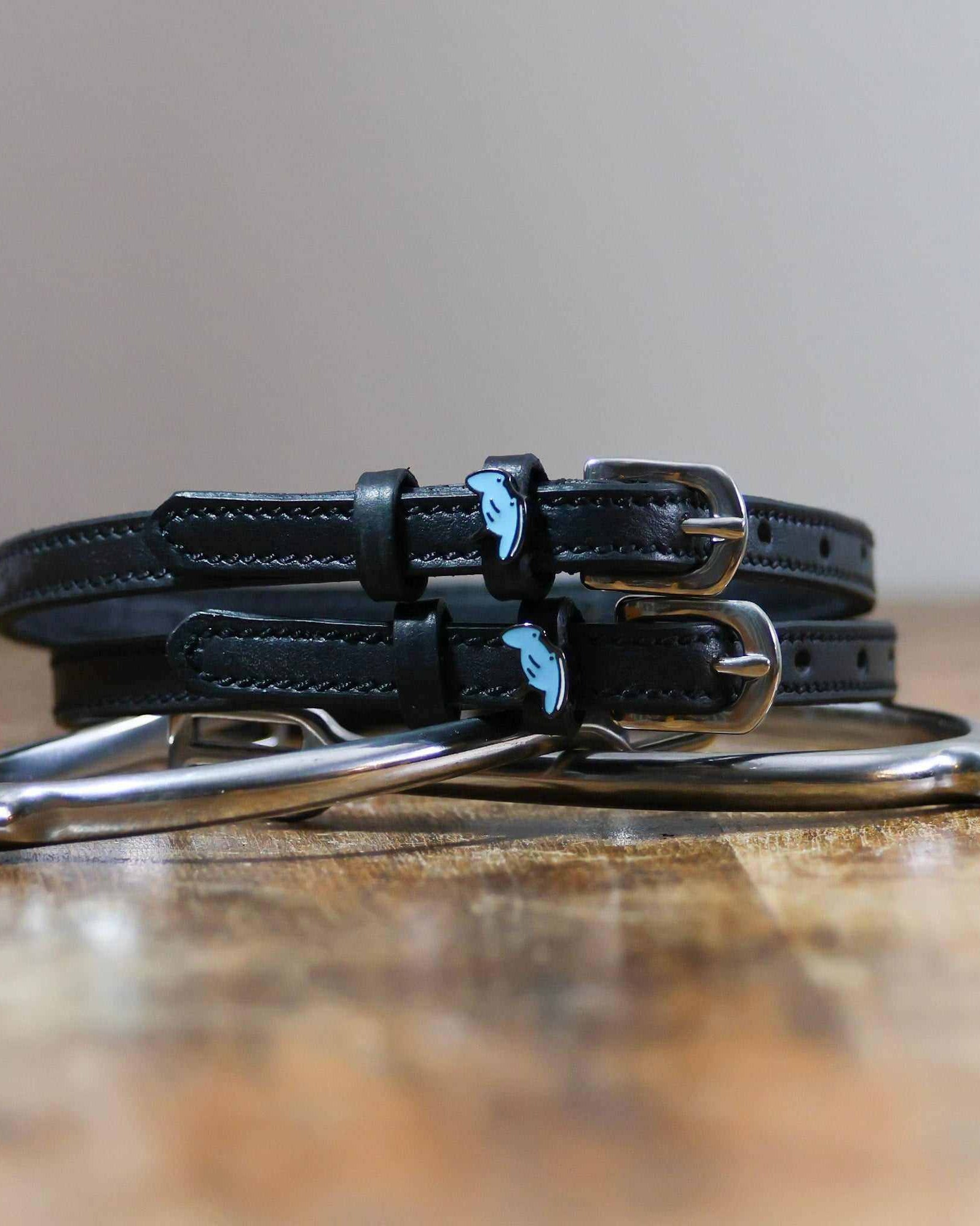 Stacked black stingray spur straps with silver buckles on wood beside a stirrup iron