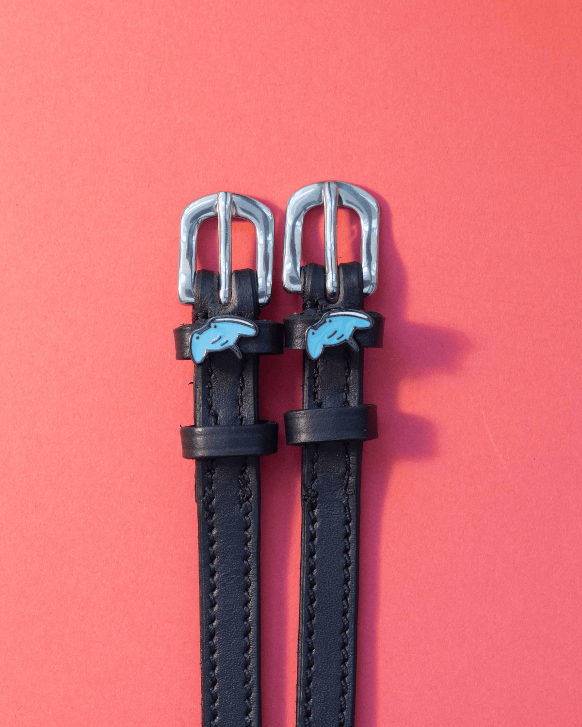 Black leather stingray spur straps with blue ray charms on a coral background, ManeJane