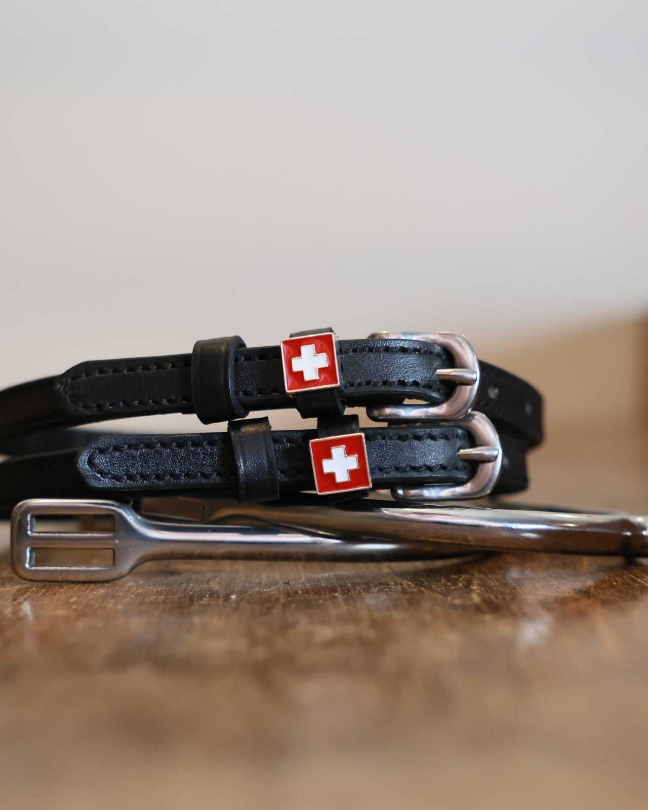Black Swiss-flag charm spur straps coiled beside a silver spur on a wood surface