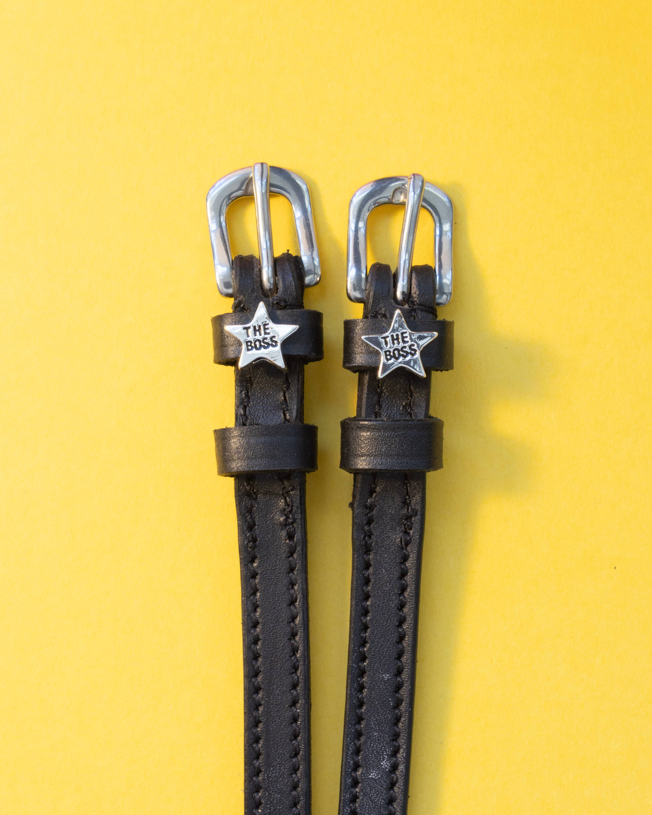 Pair of black leather Boss star spur straps flat-laid on a yellow background