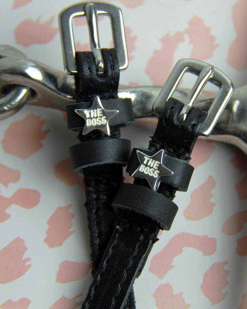Close-up of silver 'The Boss' star charms on black leather spur straps beside a metal bit