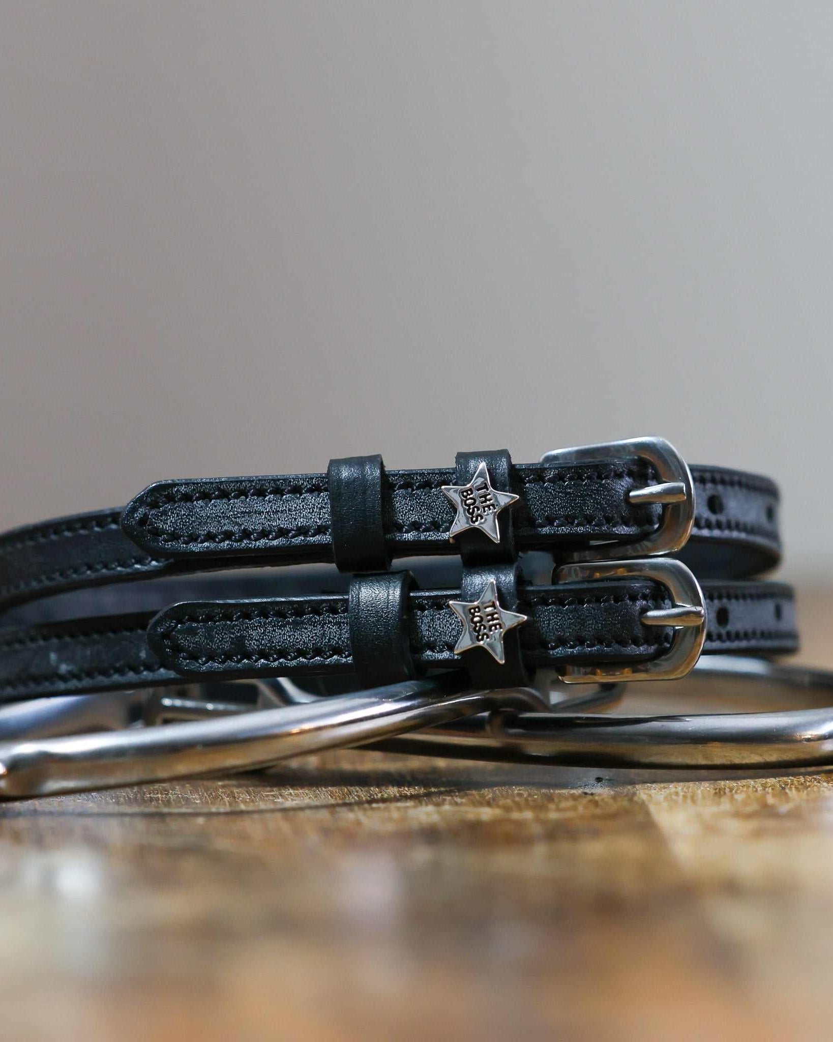 Pair of black leather Boss star spur straps coiled on wood beside a silver bit