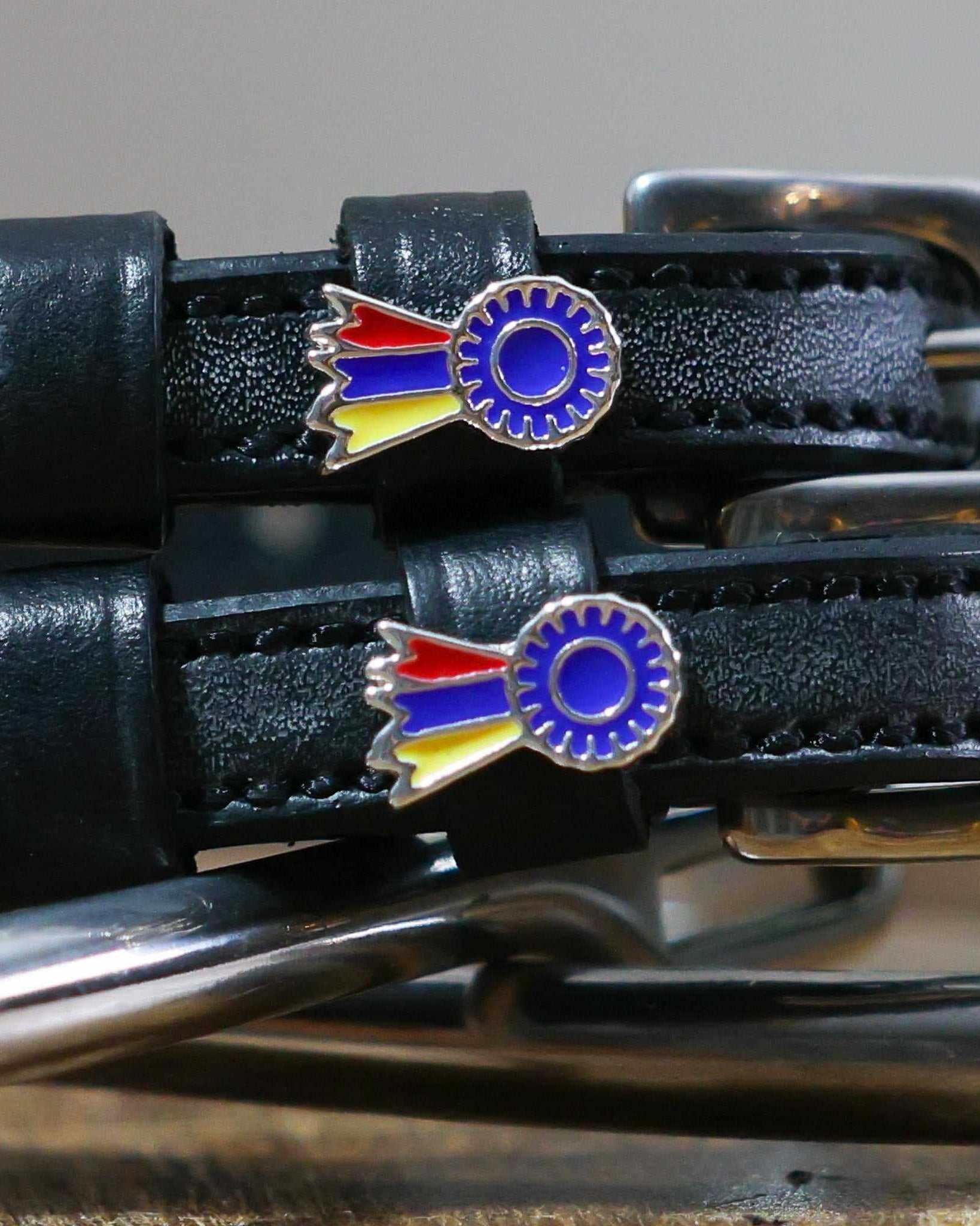 Close-up of tri-color ribbon rosette charms on black leather spur straps beside a bit
