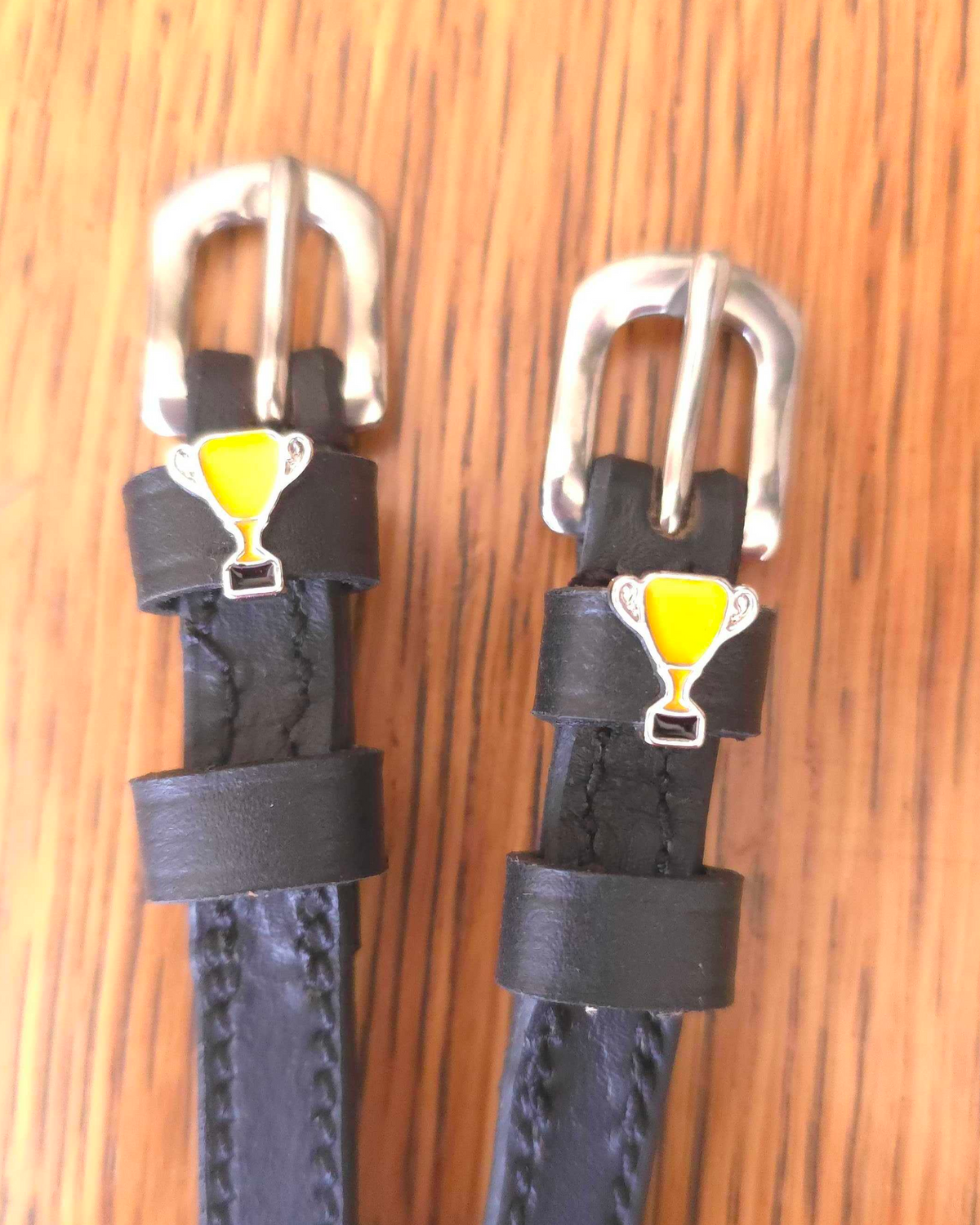 Black leather ManeJane spur straps with yellow trophy cup charms on a wood grain surface